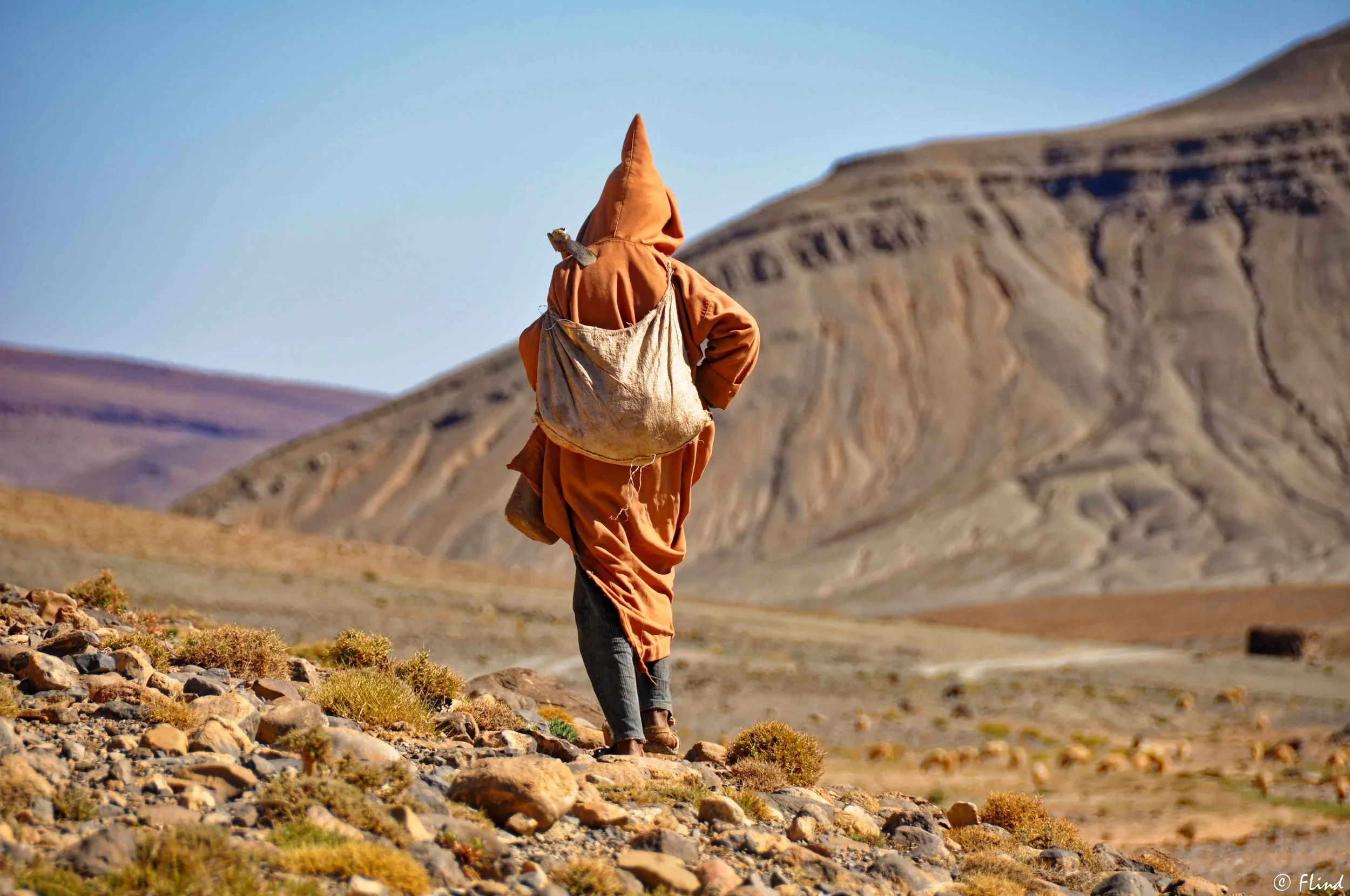 Eine Person läuft in einer trockenen, bergigen Landschaft mit wenig Vegetation, trägt einen großen Rucksack und eine Kapuzenjacke.