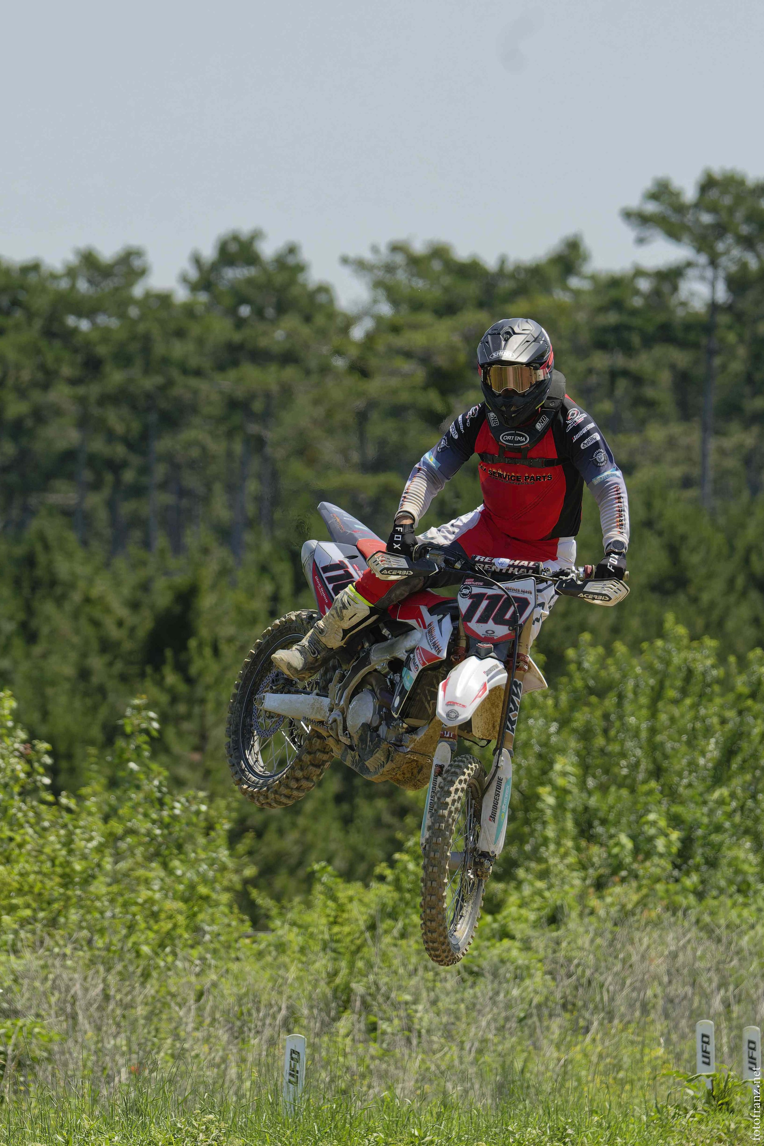 Motorradfahrer springt mit offroad-Motorrad in der Luft auf grünem Feld, umgeben von Büschen und Bäumen.