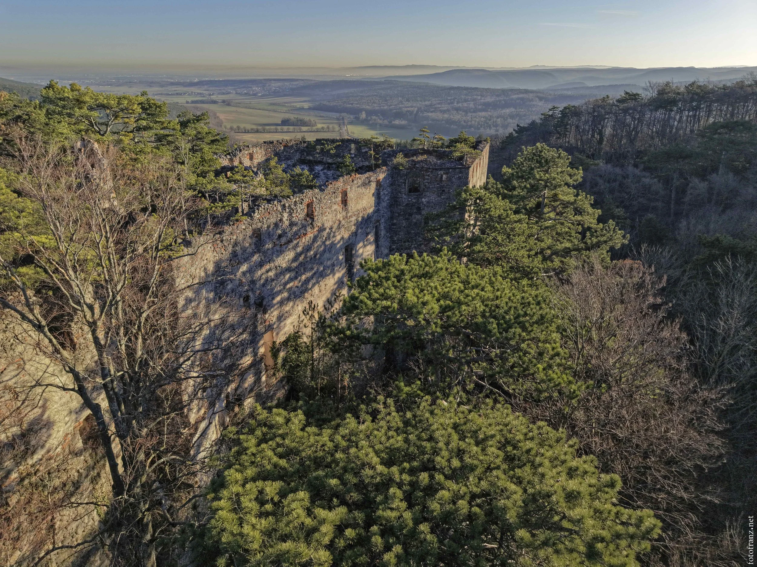 Verfallene Steinruine in einem bewaldeten Gebiet mit grünen und kahlen Bäumen, mit einer weiten Landschaft im Hintergrund.