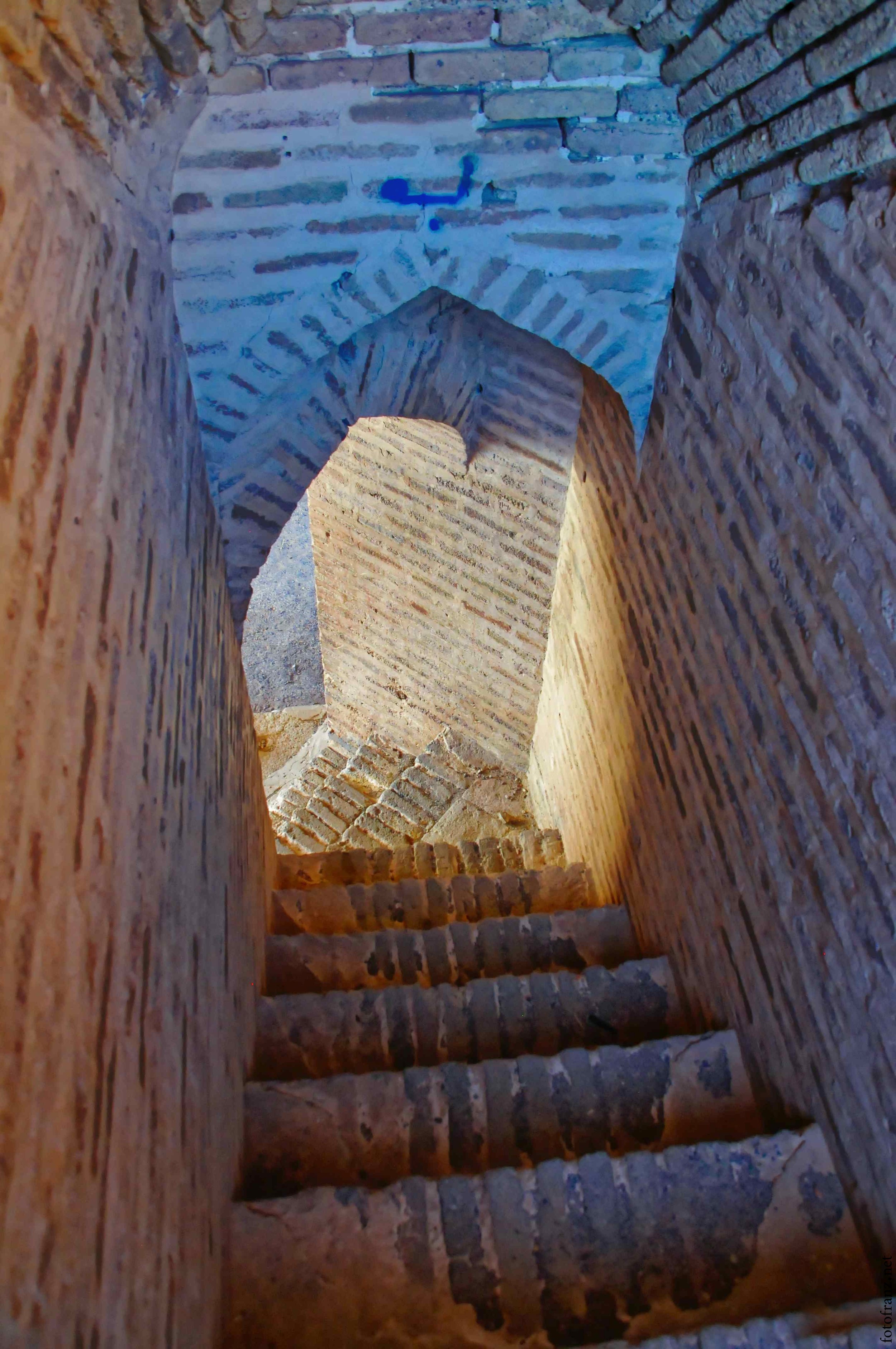 Innenansicht eines alten, steinernen Treppenhauses mit mehreren Steinstufen, umgeben von Ziegelmauern, mit Blick nach oben zur Decke und kleineren Bögen in der Architektur.