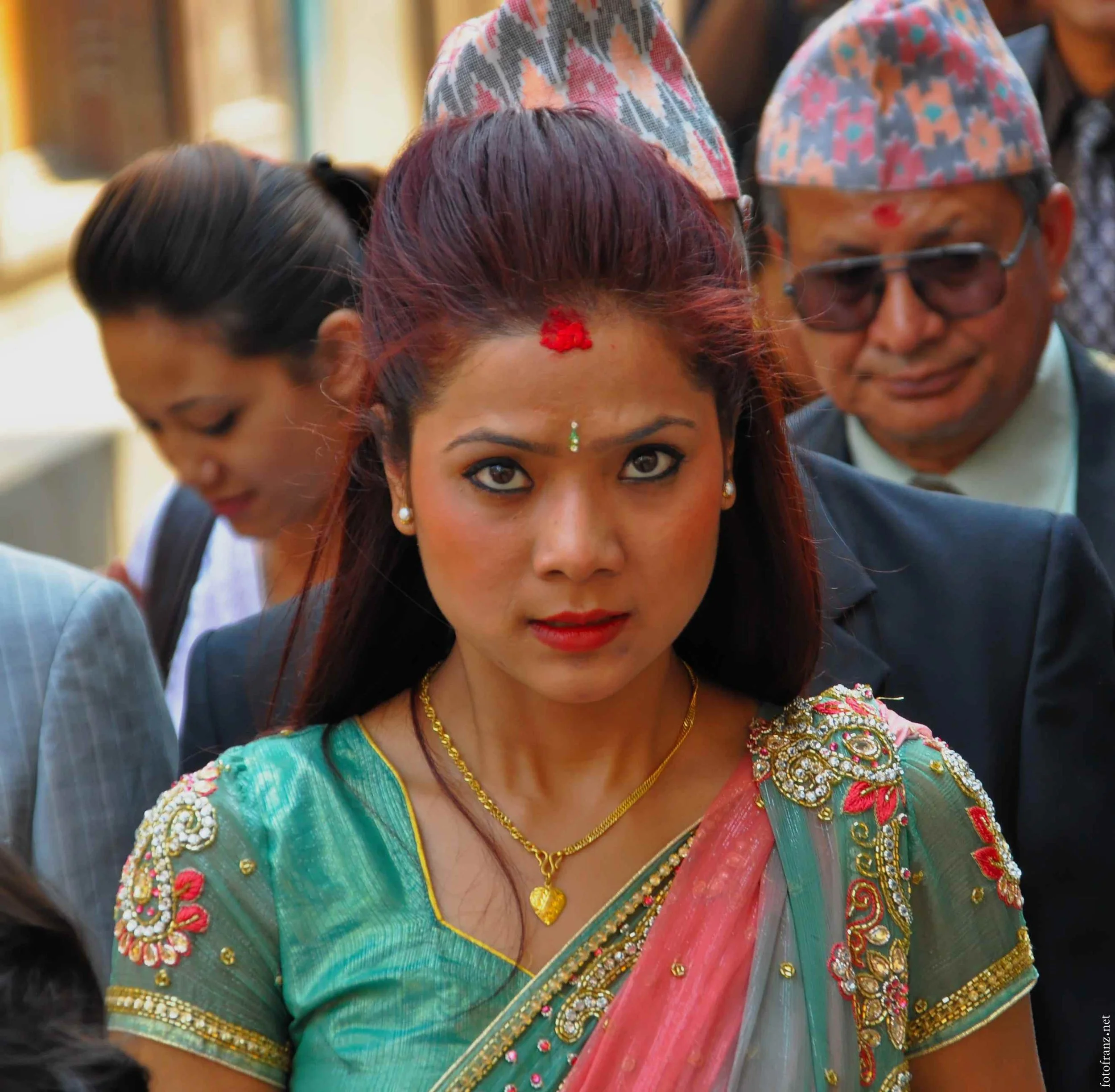 Eine junge Frau in traditioneller indischer Kleidung mit roten, goldenen und grünen Elementen, trägt Schmuck und hat einen roten Punkt auf der Stirn, umgeben von weiteren Personen in formeller Kleidung.