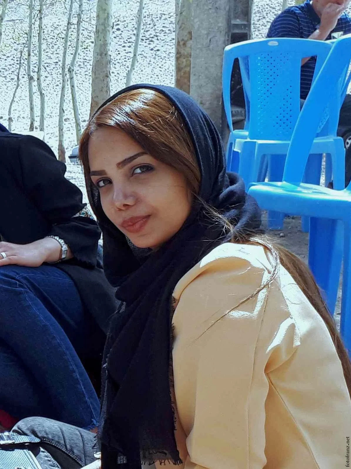 Junge Frau mit braunen Haaren, schwarzem Kopftuch und cremefarbenem Oberteil, sitzt im Freien vor blauen Stühlen.