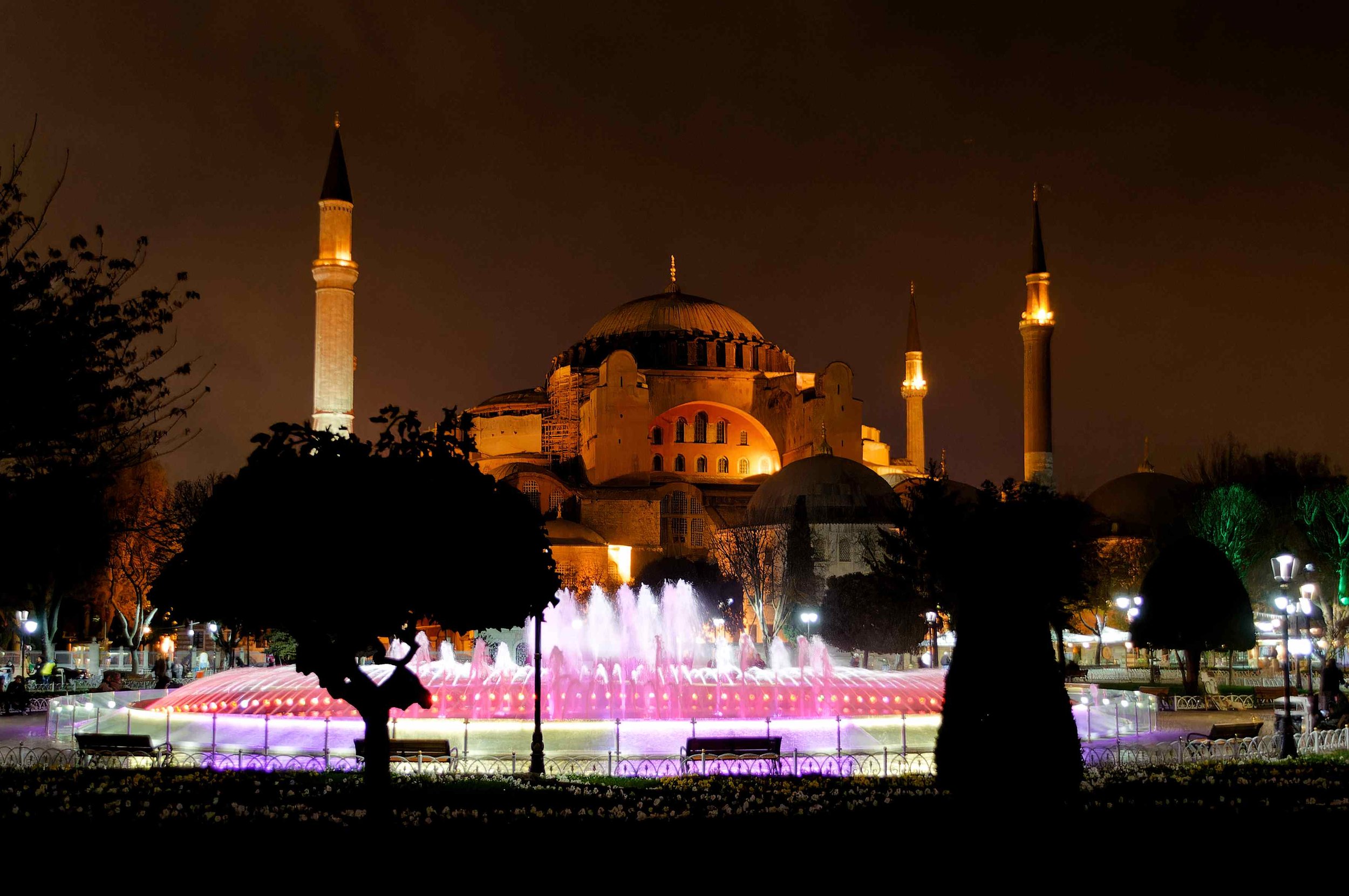 Die Hagia Sophia bei Nacht mit beleuchteten Minaretten und einem beleuchteten Springbrunnen im Vordergrund, umgeben von Bäumen und Laternen.