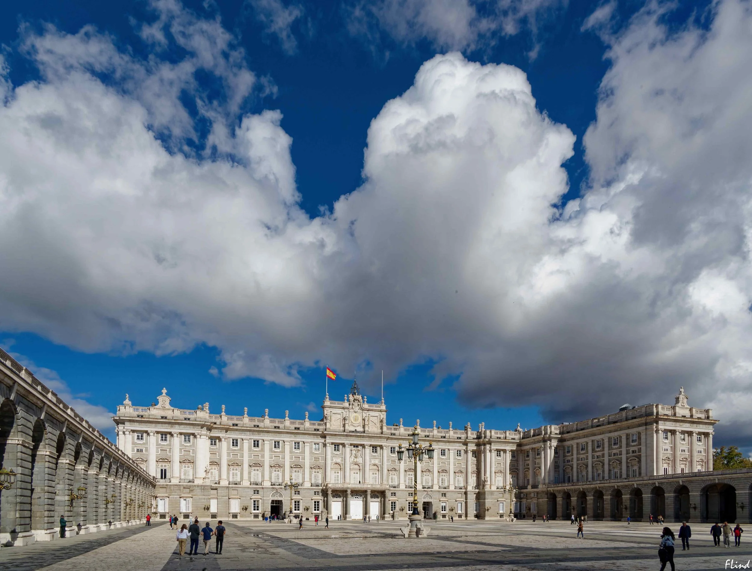 Ein prunkvolles, weißes Gebäude mit vielen Fenster und Balkonen, davor eine große, offene Plaza mit vereinzelten Menschen, darunter Kinder und Familien, unter einem Himmel mit großen, weißen Wolken.