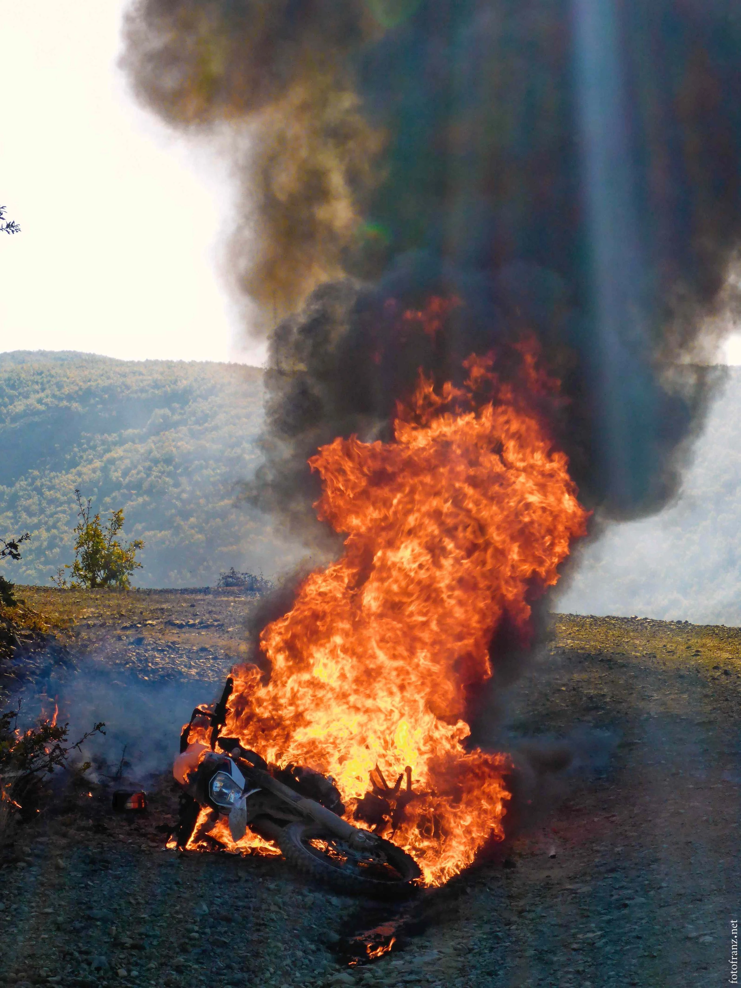 Ein brennendes Motorrad auf einer unbefestigten Straße mit dunklem Rauch aufsteigend in die Luft.