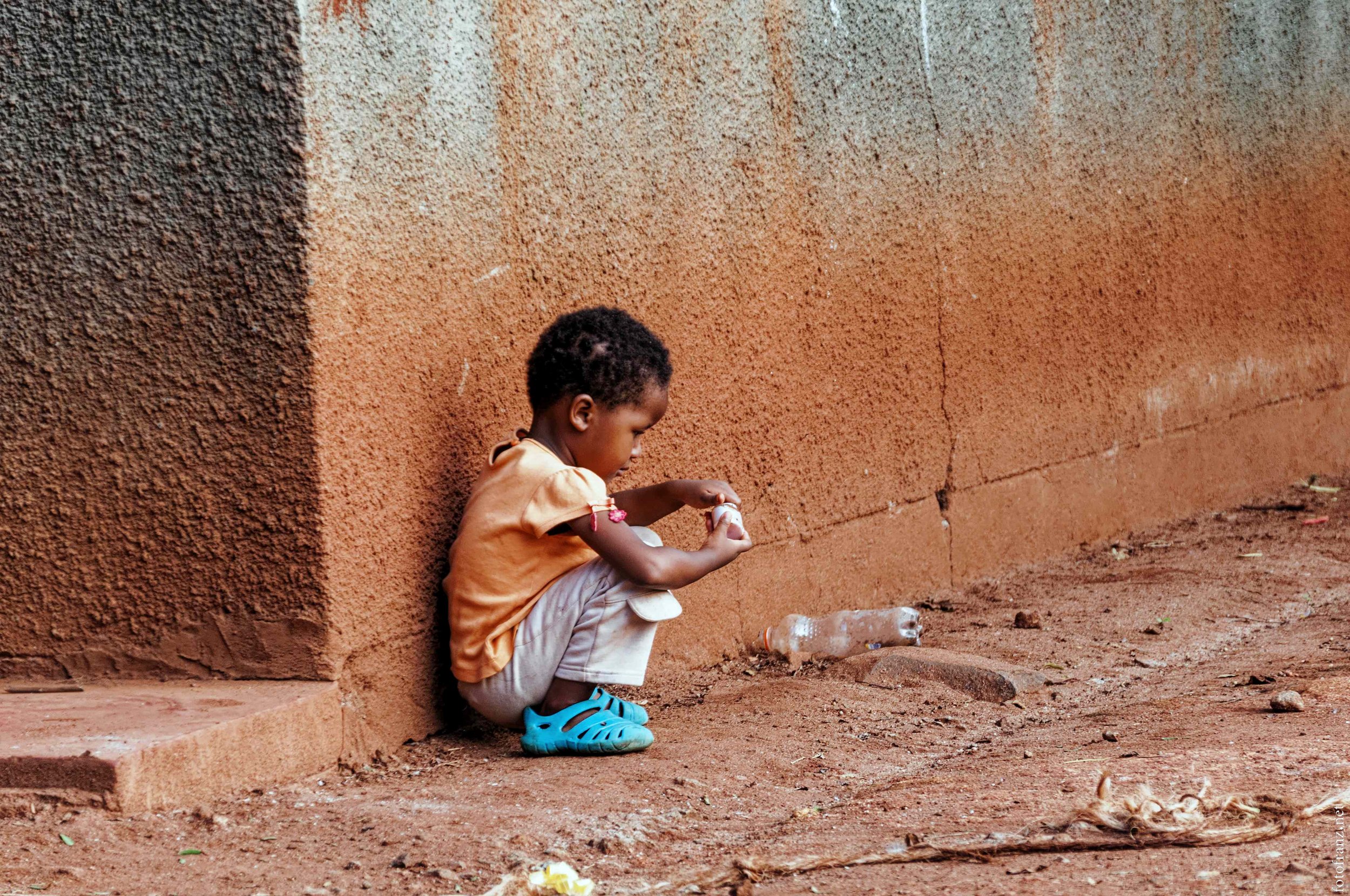Ein Junge sitzt auf dem Boden an einer braunen Wand und spielt mit einer kleinen Figur, neben einem leeren Plastikflaschen.
