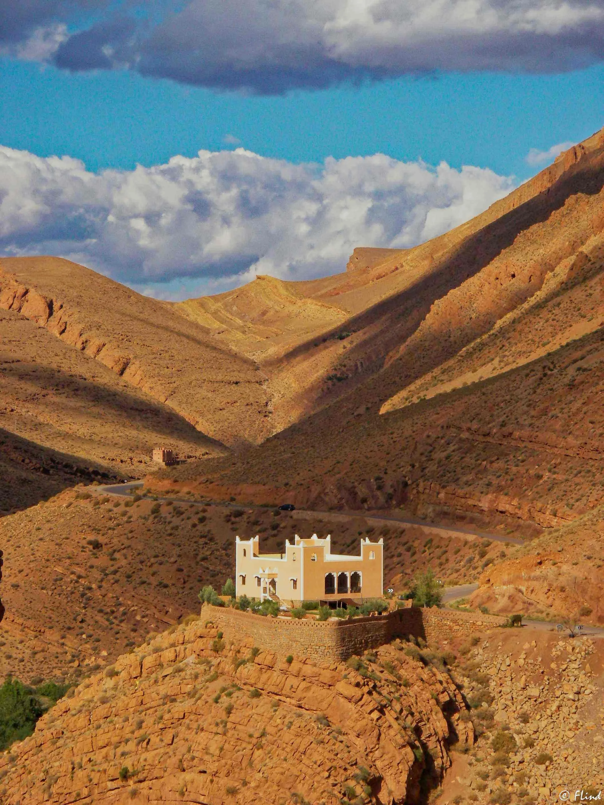Ein Haus auf einer Klippe in einer menschenleeren, bergigen Wüstenlandschaft unter einem bewölkten Himmel.