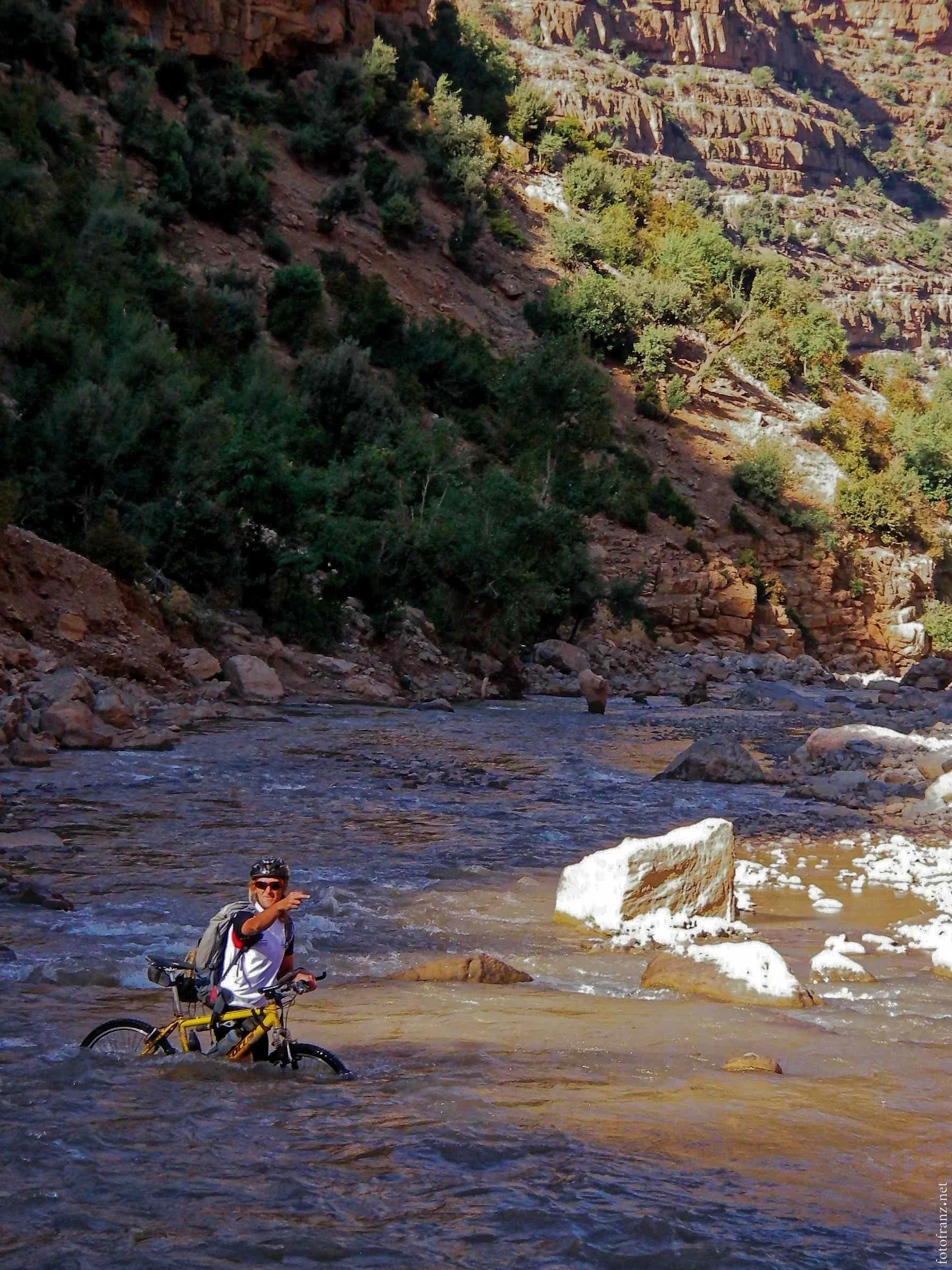 Ein Mountainbike-Fahrer steht im Fluss mit Bergen im Hintergrund, zeigt auf die Kamera, trägt Helm und Rucksack.