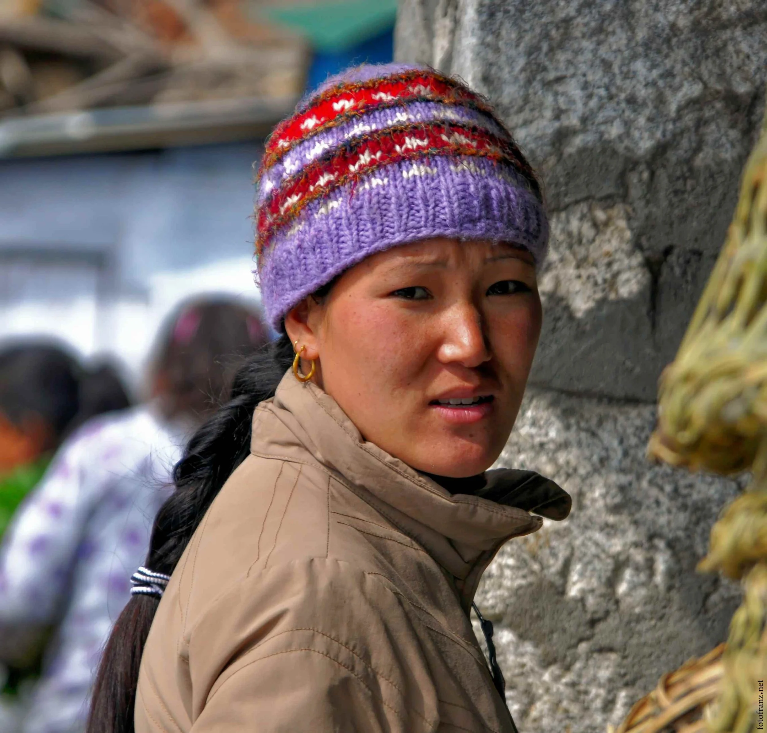 Naheaufnahme eines jungen Mädchens mit dunklem Haar, das eine bunte Wollmütze trägt, während es an einer Steinmauer steht.
