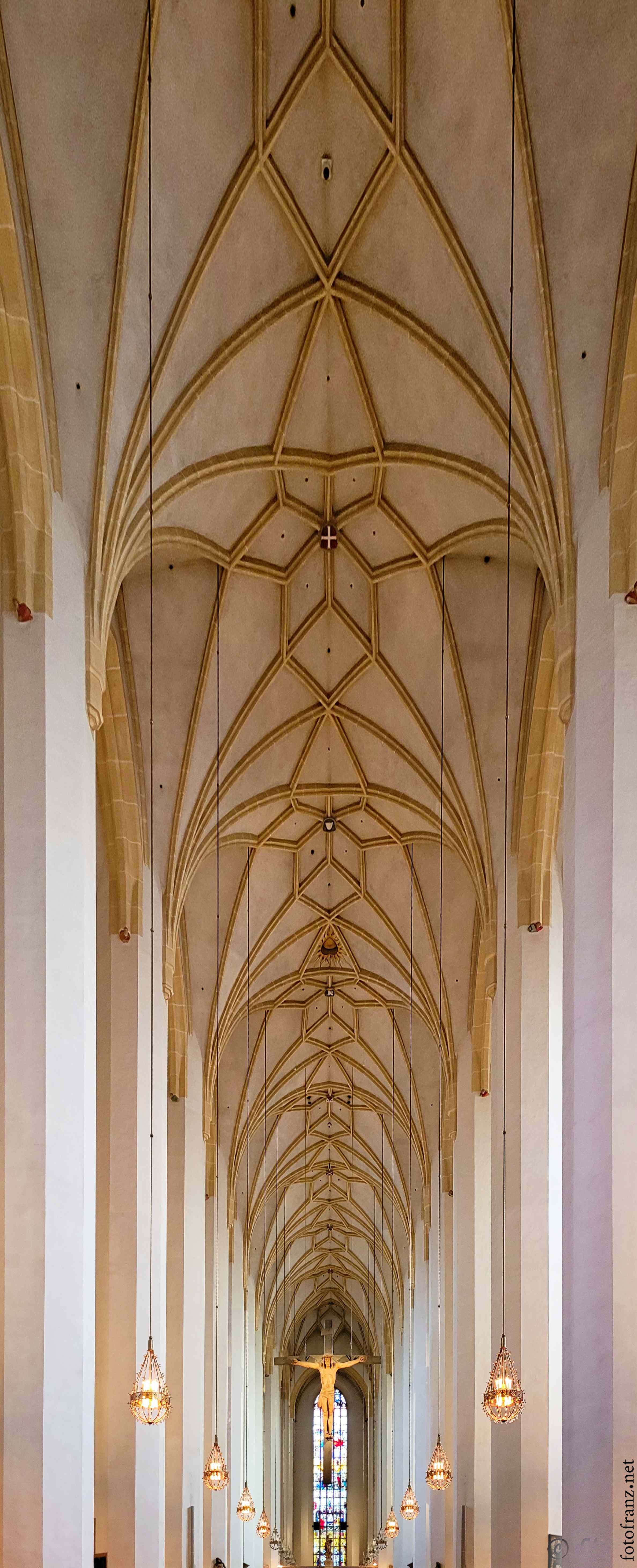 Innenansicht einer Kirche mit hohen Gewölbedecken, einer hölzernen Kreuze und einer bunten Fenster im Altarbereich.