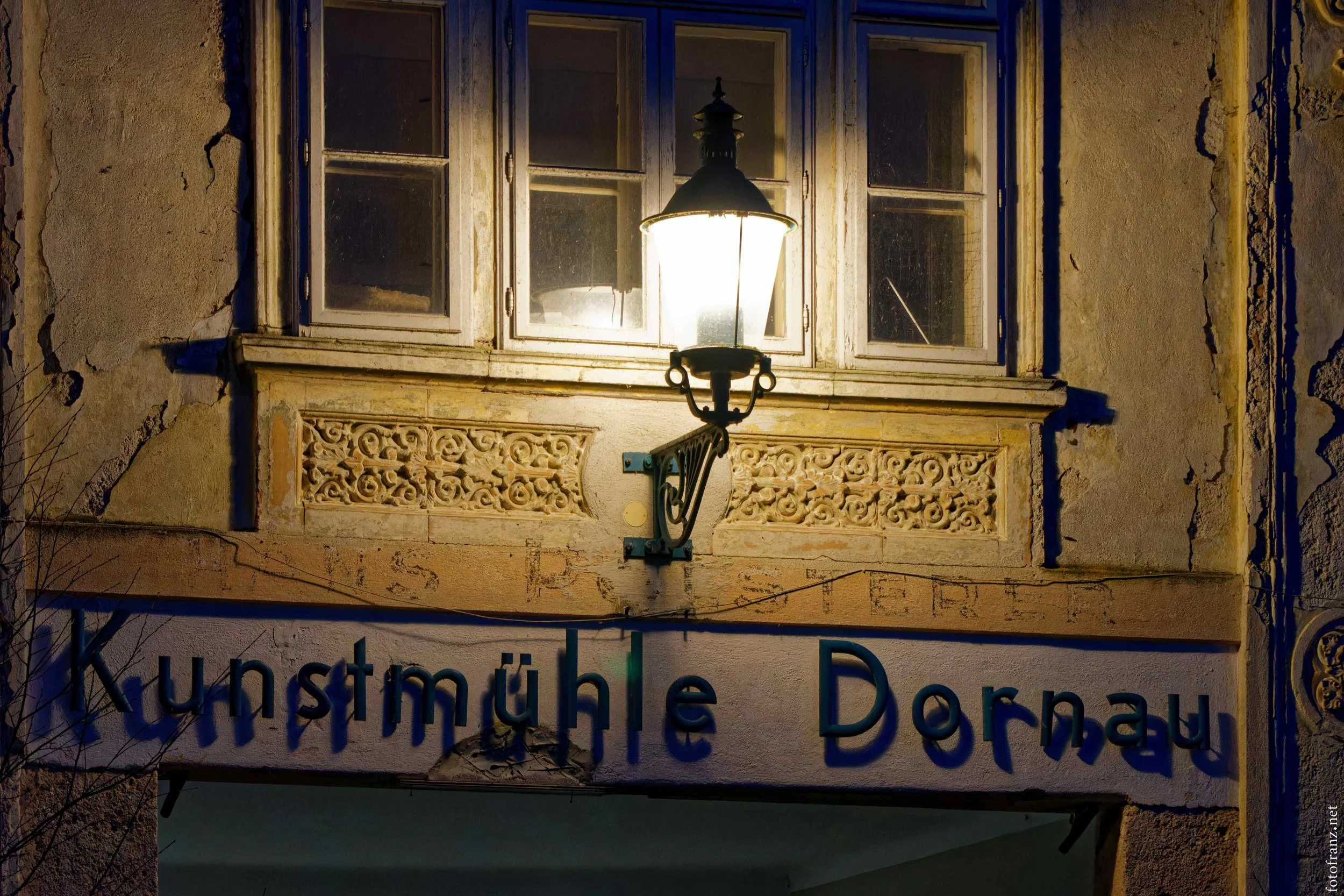 Ein beleuchteter Laternenstrahler an einer alten Hausfassade mit einem Fenster und der Aufschrift "Kunstmühle Dornaub"