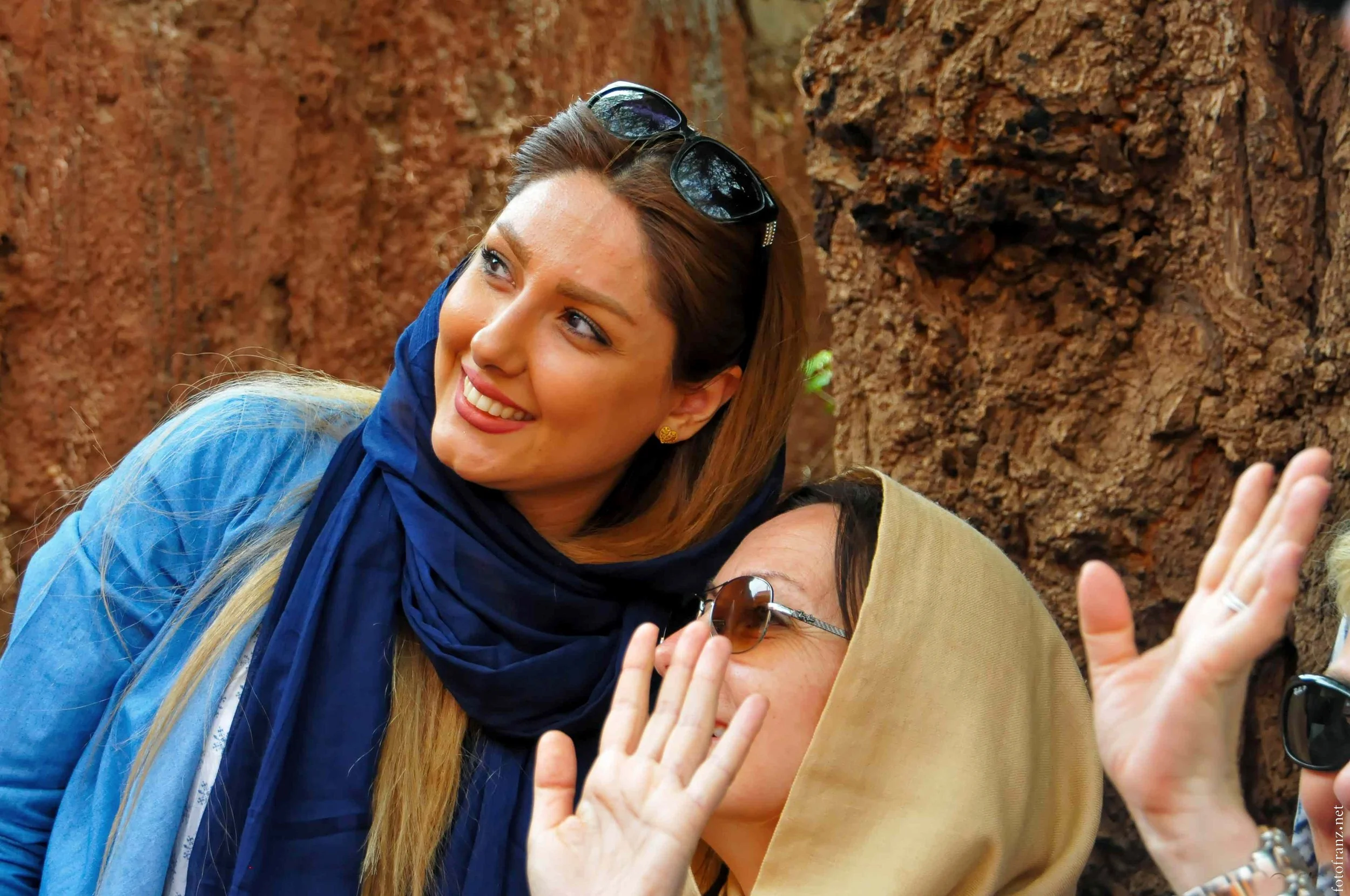 Zwei lachende Menschen beim Gespräch, einer trägt eine beige Kopfbedeckung und Brille, die andere Sonnenbrille auf dem Kopf, vor einem Felsen.