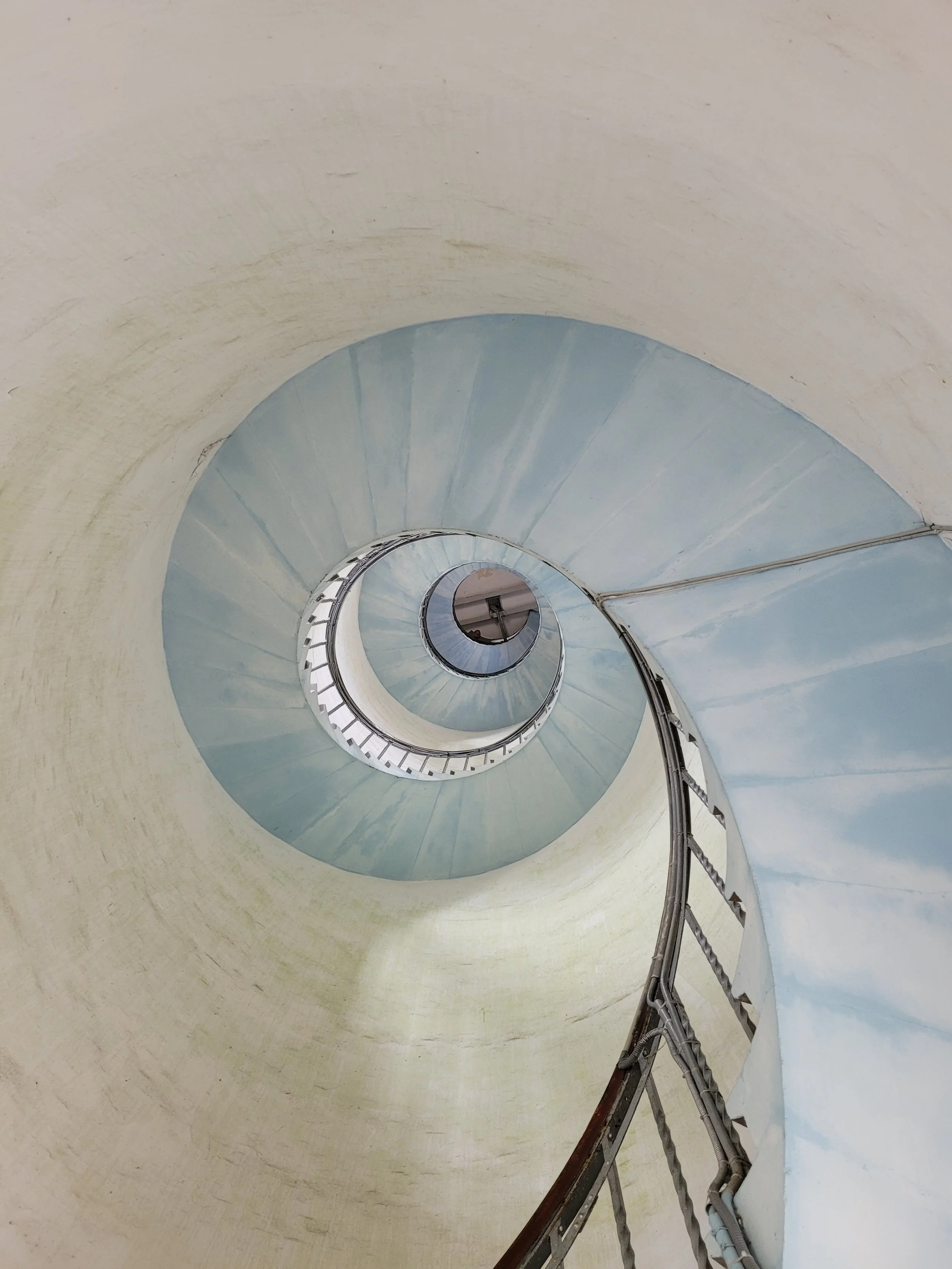 Innenansicht einer spiralförmigen Treppe, die nach unten führt, mit blauer Wandbemalung und gelblicher Decke, Blick nach unten in den Abstieg.