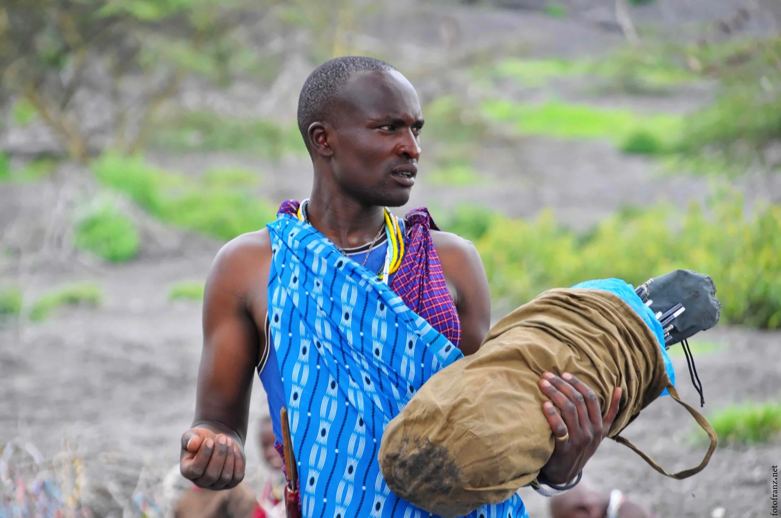 Mensch in traditioneller Kleidung hält gepackten Rucksack in der Hand im Freien, vermutlich bei einer Reise oder einer Zeremonie.