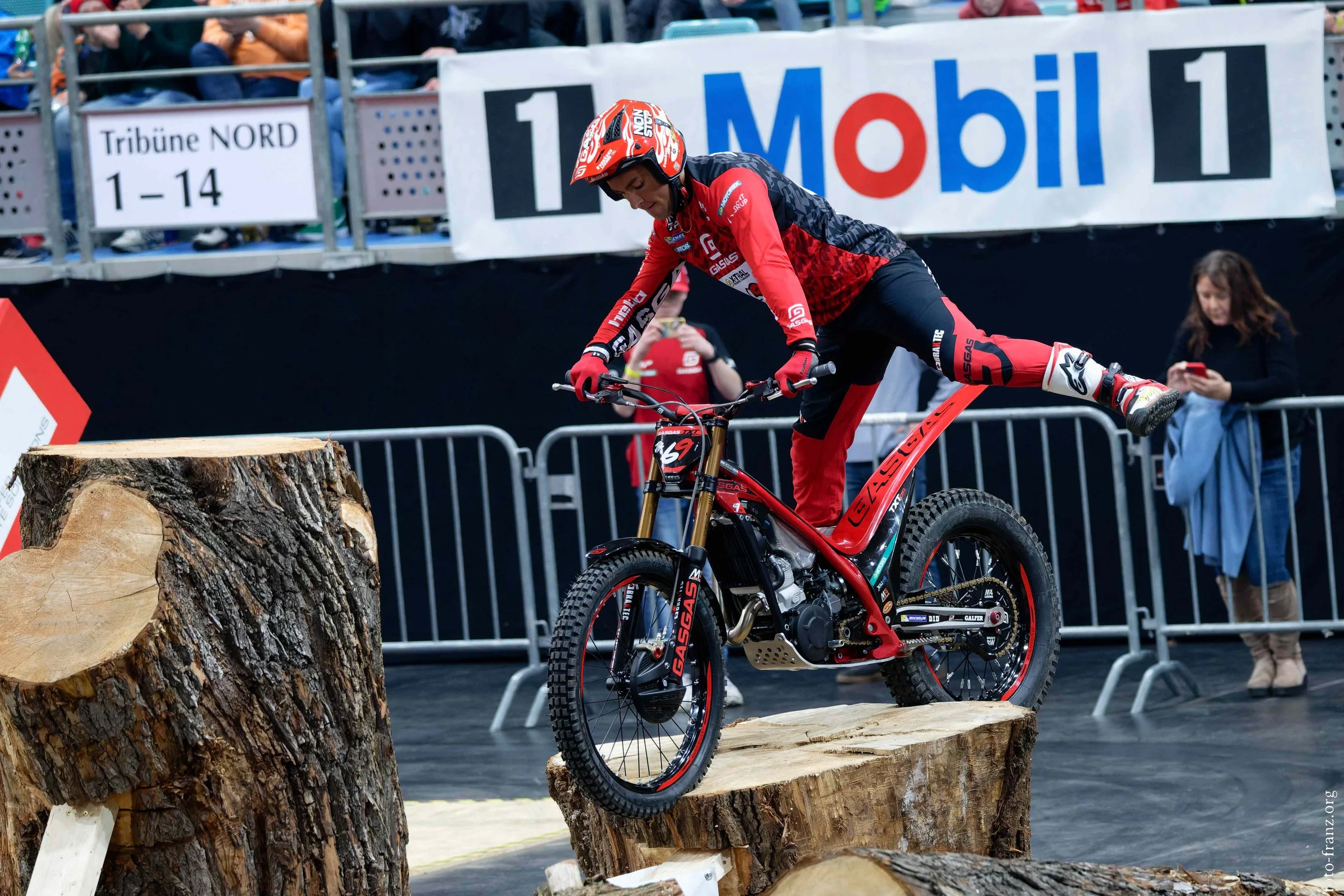 Ein männlicher Mountainbike-Fahrer in Rot und Schwarz bei einem Stunt im Wettkampf, springt auf einen Holzstamm, im Hintergrund sind Zuschauer und Werbebanner.