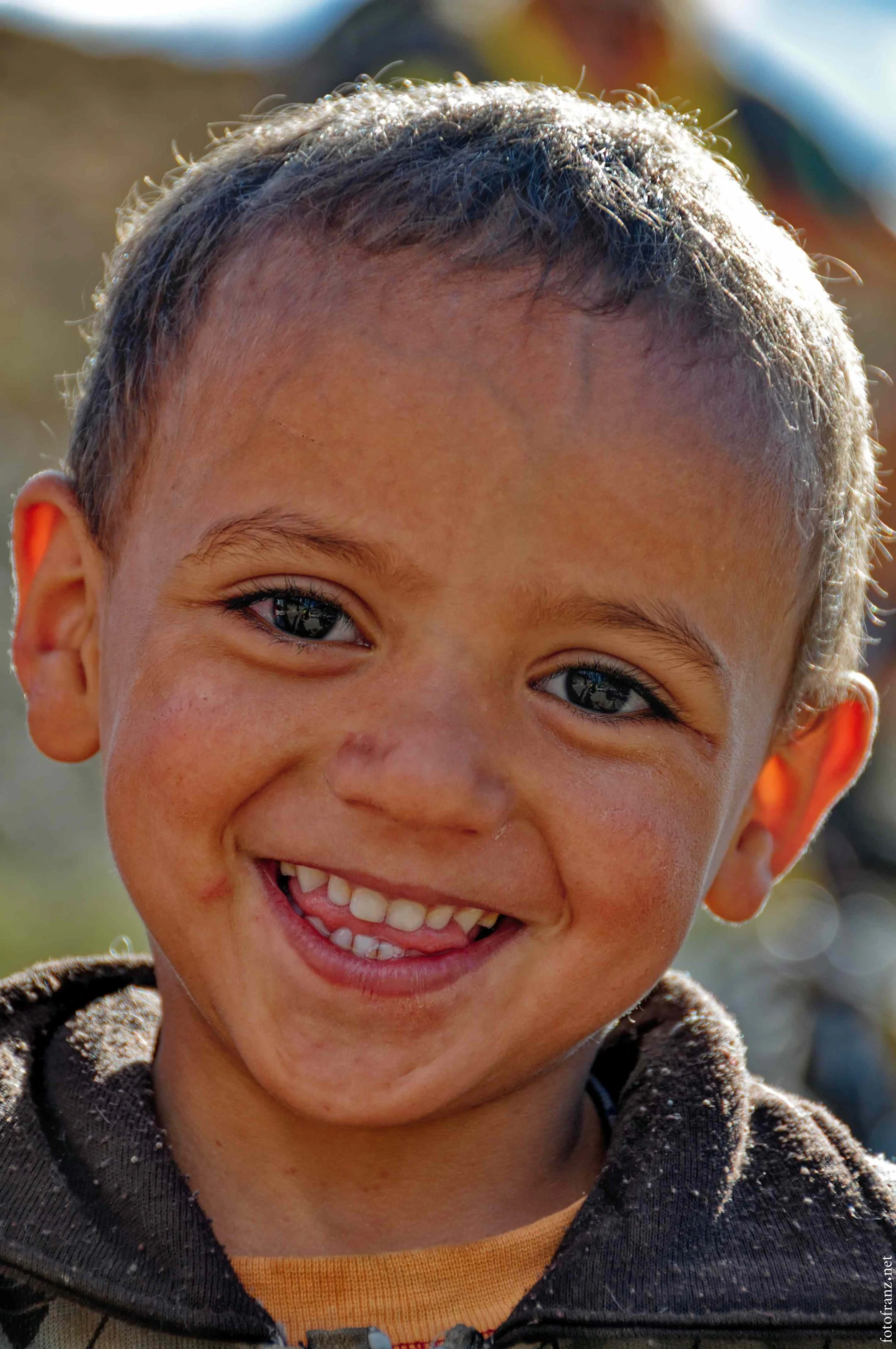 Lächelnder Junge mit blauen Augen, kurze braune Haare, verschmutztem Gesicht und Kleidung, im Freien bei Sonnenlicht.