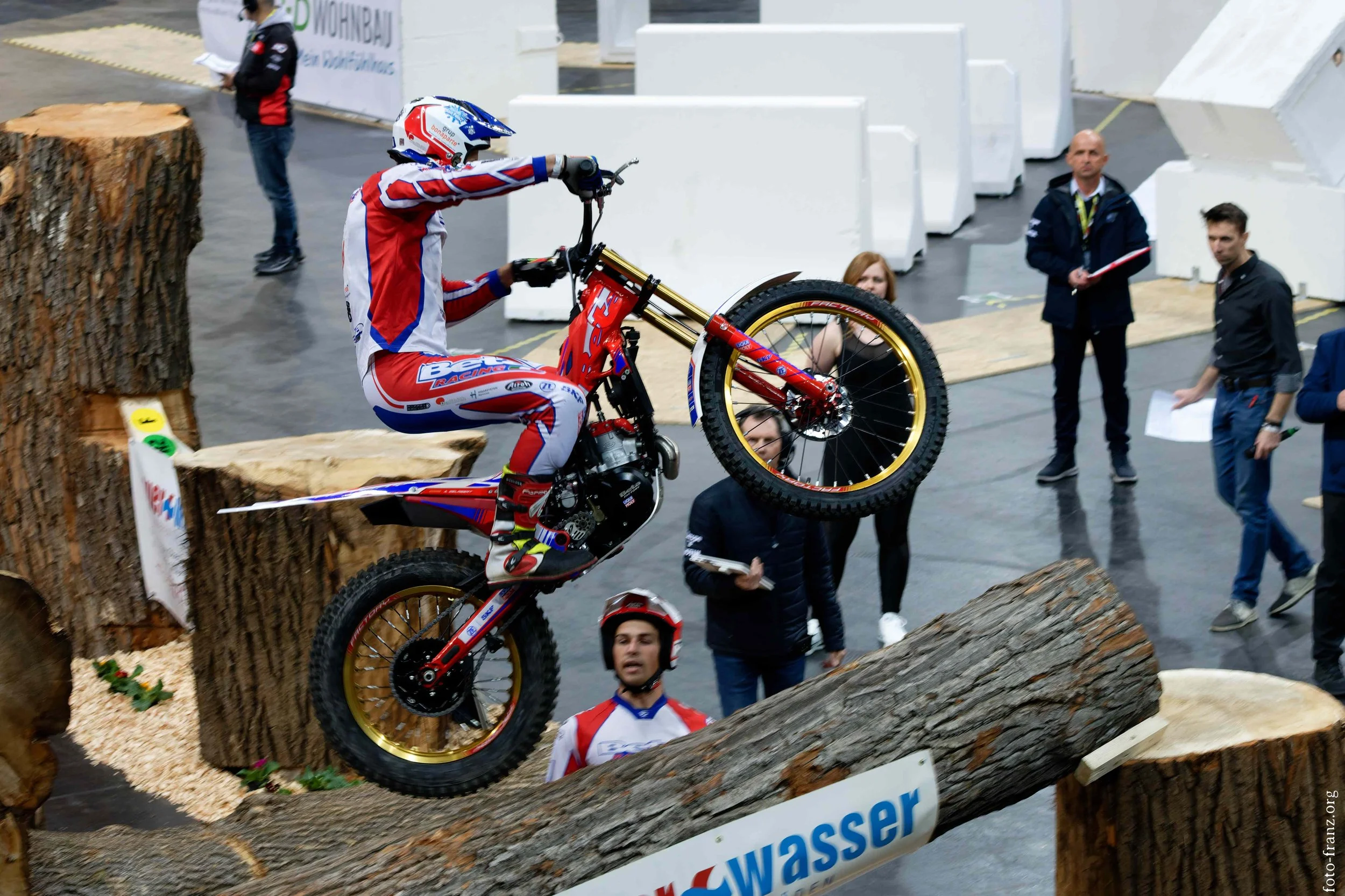 Motorradfahrer bei einer Stunt-Show auf Holzstämmen, umgeben von Zuschauern und Verantwortlichen.