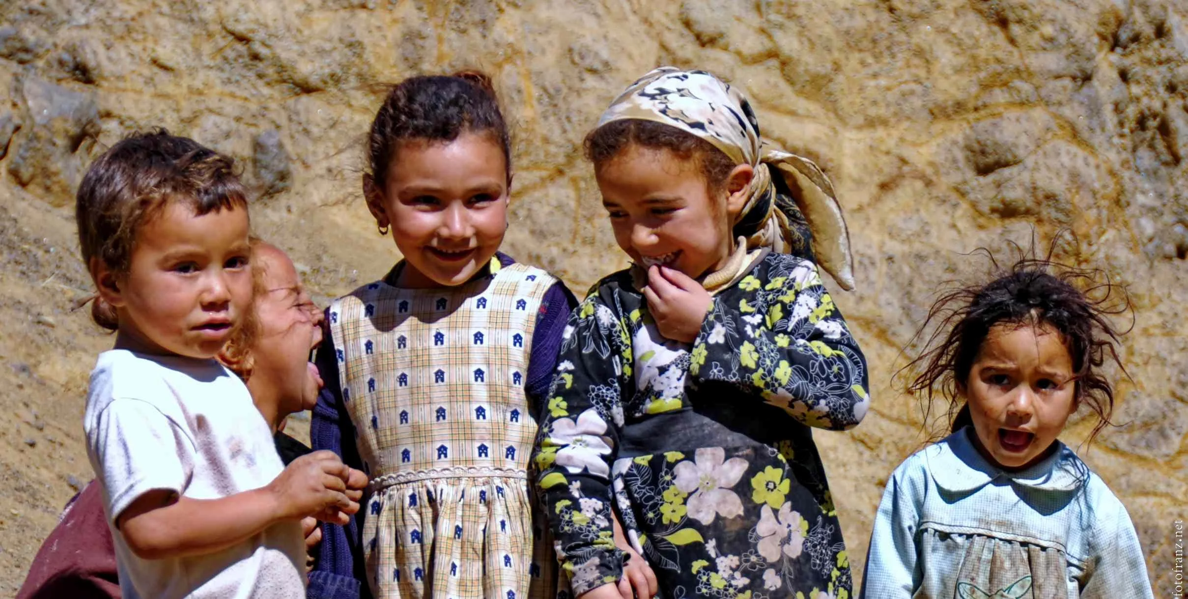 Fünf Kinder, die vor einer Felswand stehen, lachen und sprechen miteinander, tragen bunte Kleider.