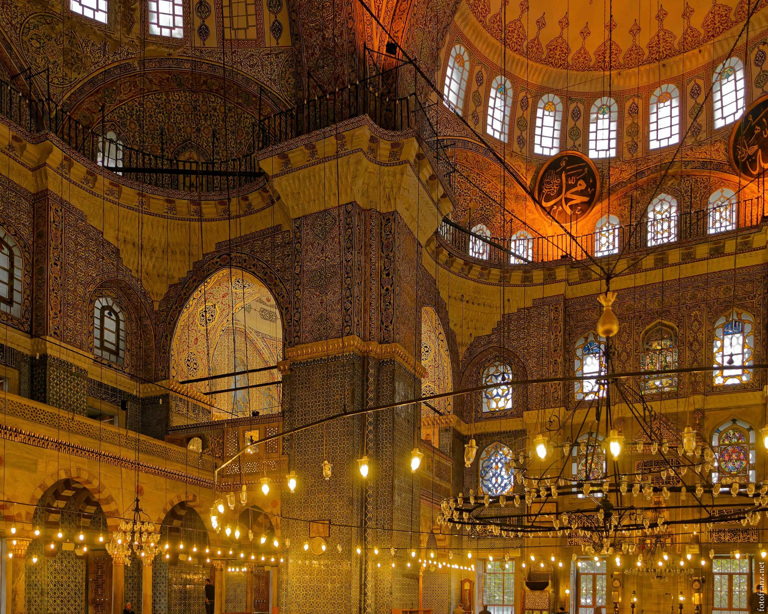 Innenansicht einer prächtigen Moschee mit vieler Ornamentierung, buntenFenstern und großen goldenen Kronleuchtern.