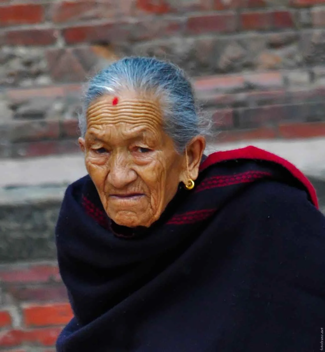 Eine alte Frau mit grauem Haar, trägt ein schwarzes Kleid und eine rote Schleife am Kopf, steht vor einer Backsteinmauer.