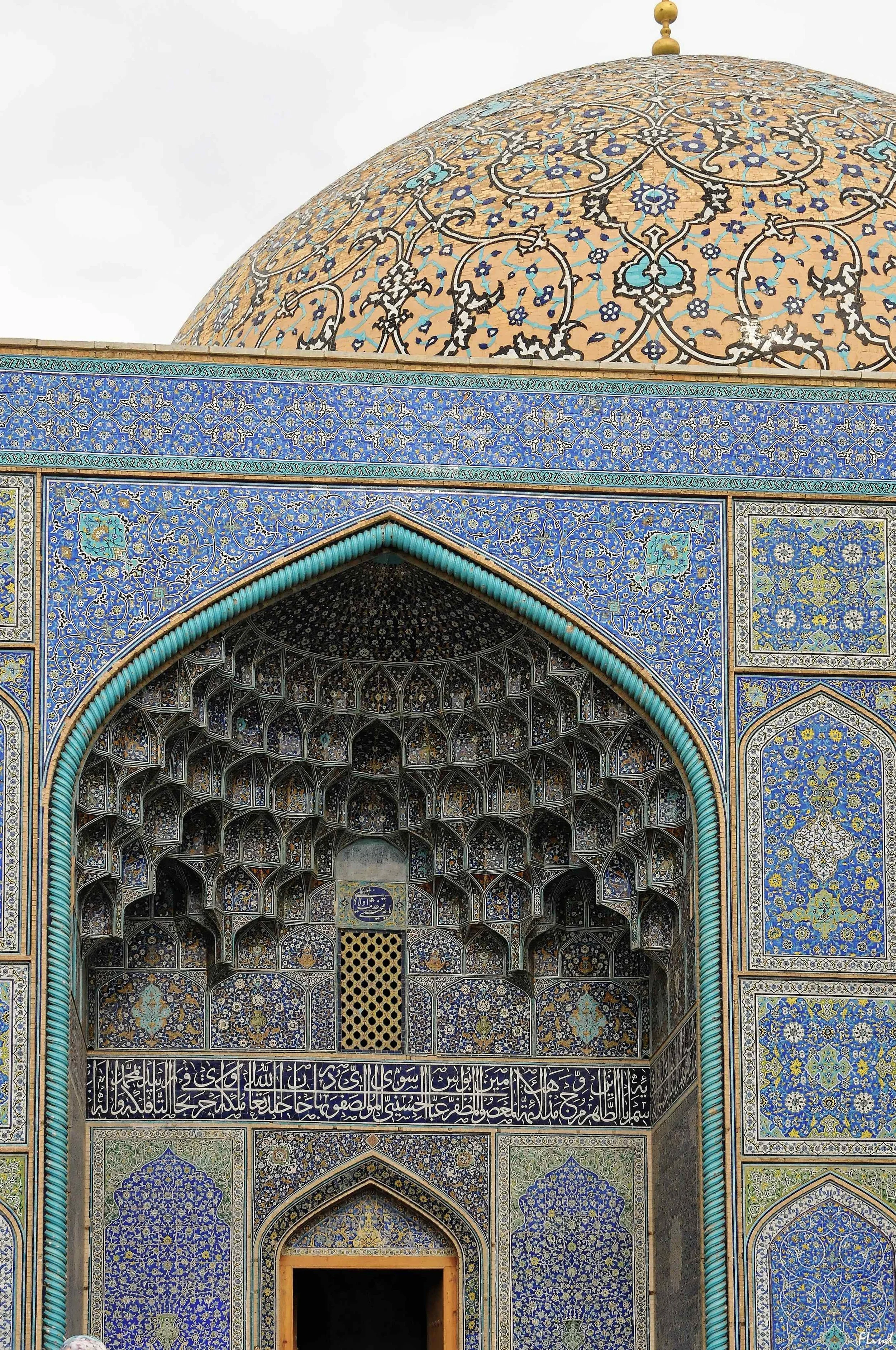 Nahaufnahme der kunstvoll verzierten Fassade eines islamischen Gebäudes mit blauen und goldenen Mosaiken, einer Kuppel und einem durchbrochenen Gitterfenster.