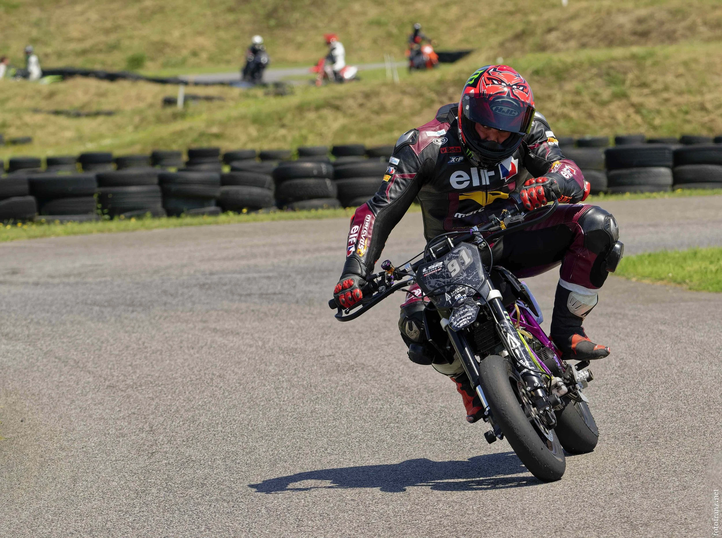 Motorradfahrer in Rennkleidung bei einer Kurvenfahrt auf einer Rennstrecke, im Hintergrund Reifenstapel und andere Fahrer.