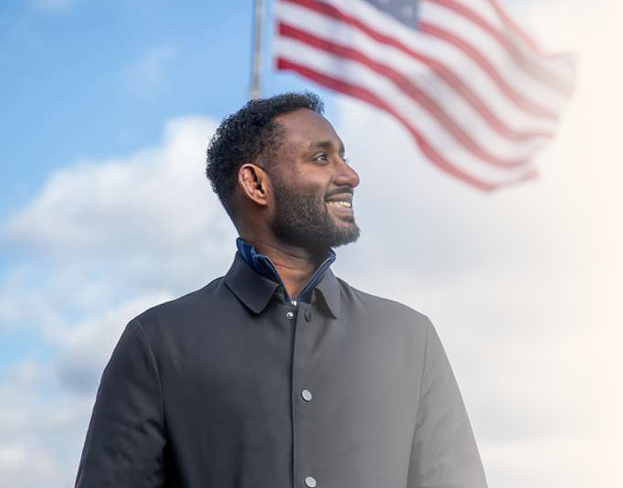 Man in a black jacket standing outdoors near an American flag, with a blue sky and clouds in the background.