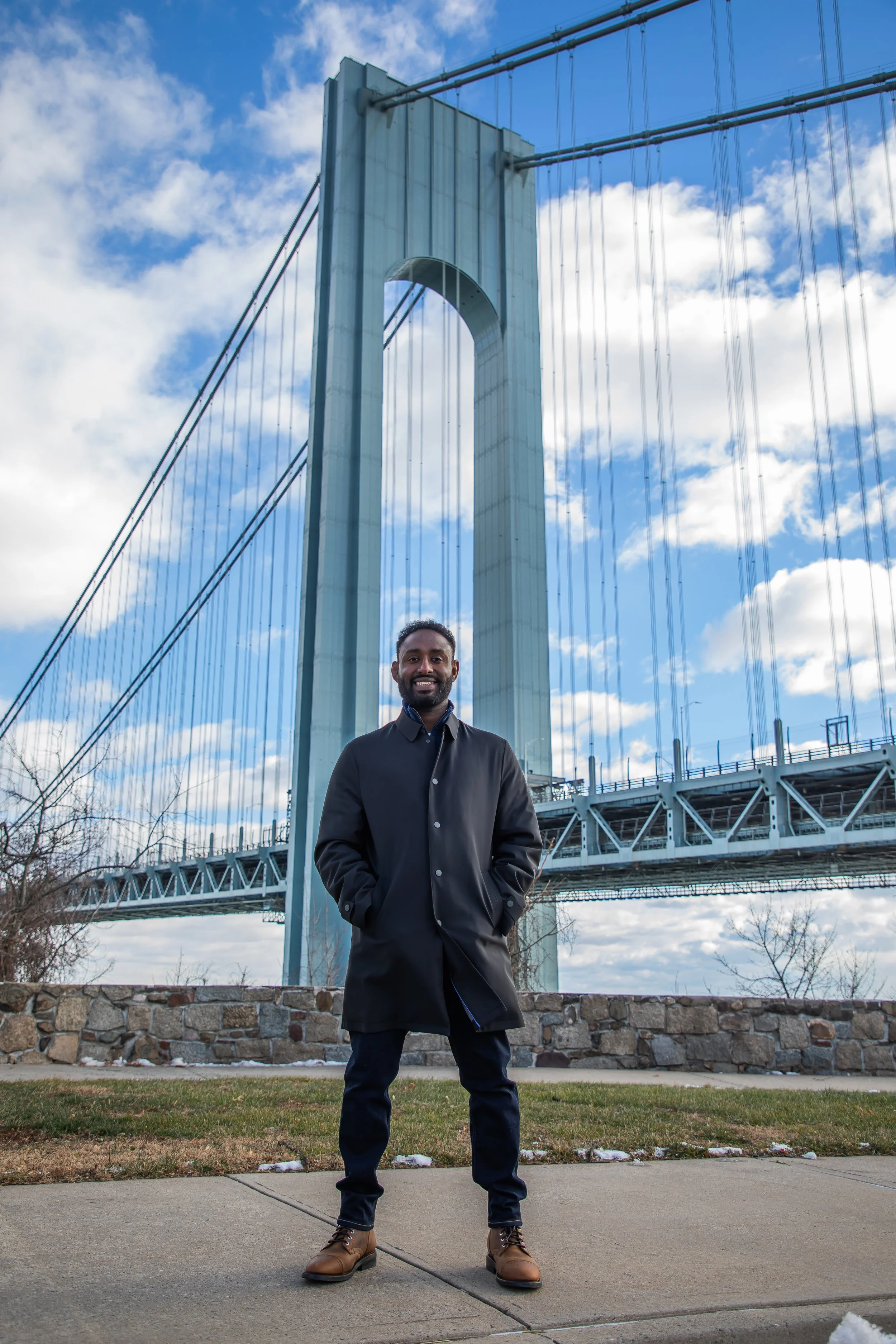 A smiling man wearing a black coat, dark jeans, and brown boots standing on a sidewalk in front of the Brooklyn Bridge with a partly cloudy sky in the background.
