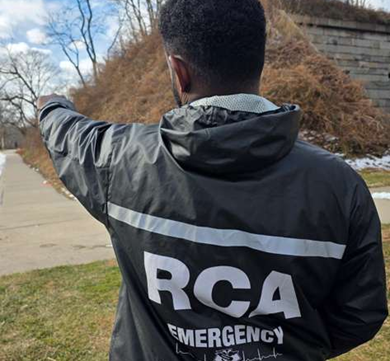 A person wearing a black jacket with reflective stripes and the words 'RCA Emergency' on the back, standing outdoors pointing to the left. Tree line and sidewalk visible, some snow patches present.