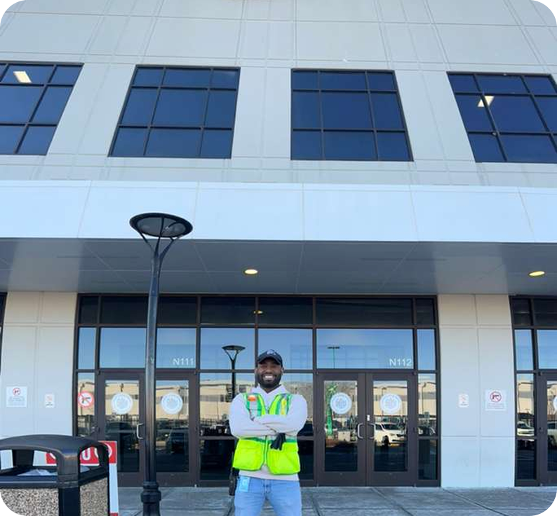 Man standing outside a modern building with large glass windows, wearing a yellow safety vest and a cap, smiling with arms crossed.