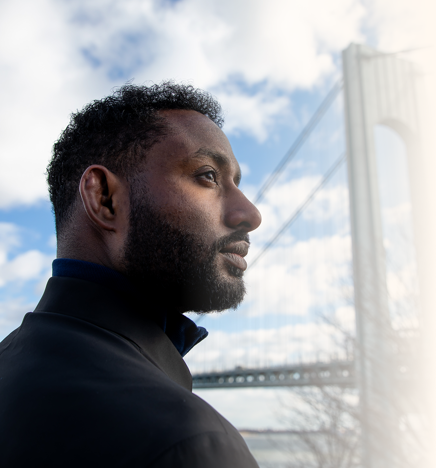Side profile of a man with beard looking into the distance with a bridge in the background.