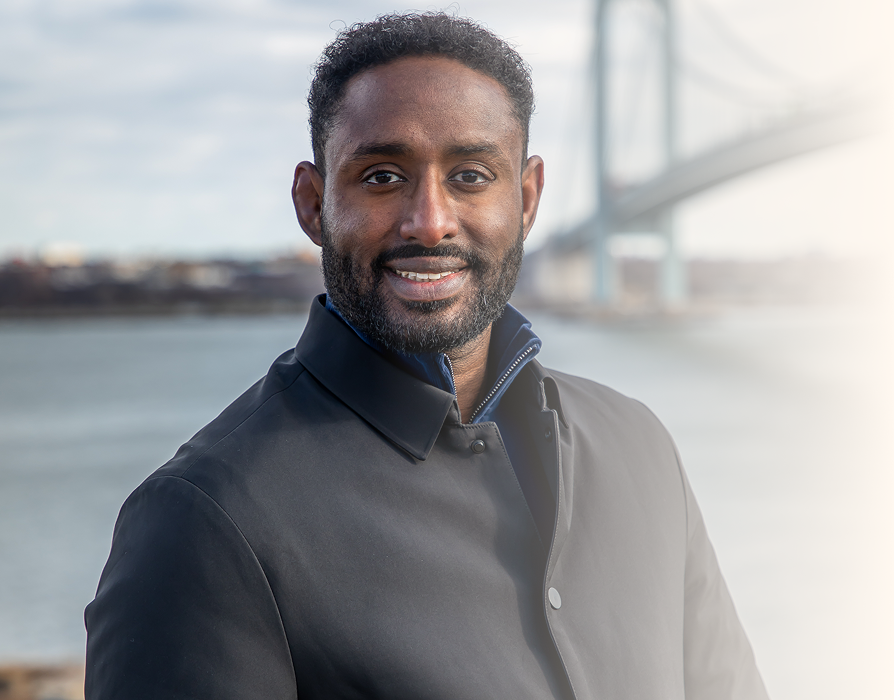 A smiling Black man with short curly hair and a beard, wearing a black jacket, standing outdoors near a body of water with a bridge in the background.