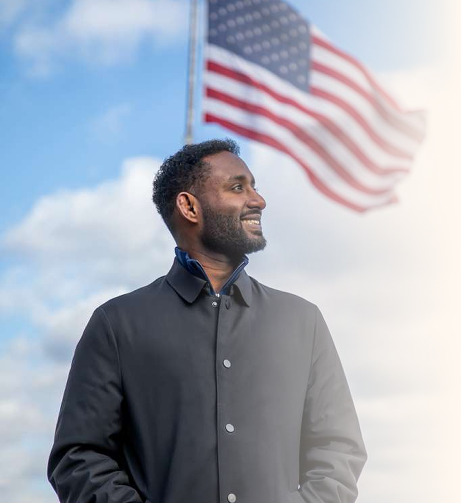 A man standing outdoors with an American flag waving behind him.