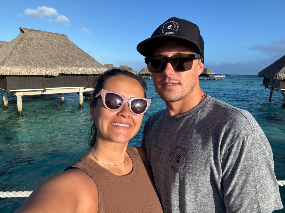 A smiling woman and man wearing sunglasses taking a selfie at a tropical overwater bungalow resort, with turquoise water and thatched-roof overwater villas in the background.