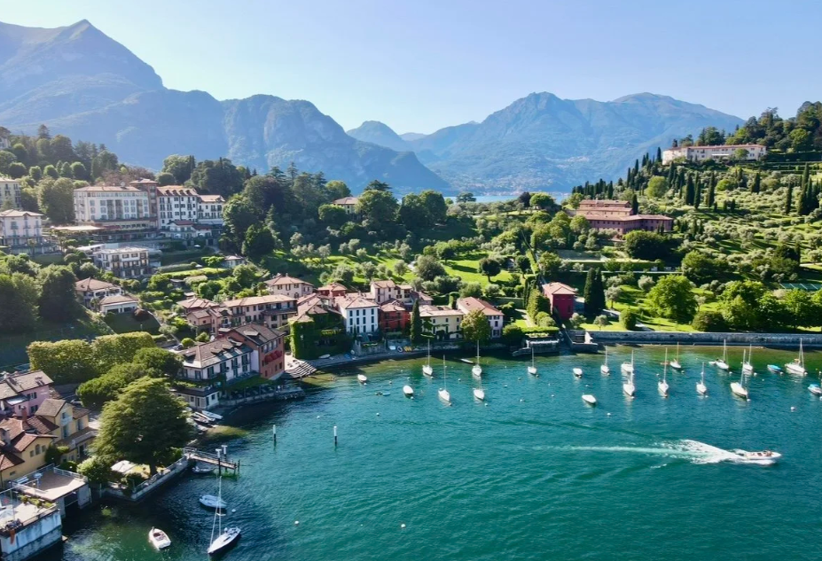 A lakeside town with colorful houses, surrounded by lush green hills and mountains in the background. Sailboats are docked on the water, and the scene is bright and sunny.