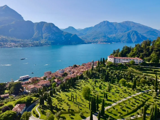A scenic view of a lake surrounded by mountains, with a town and a lush green hillside in the foreground.
