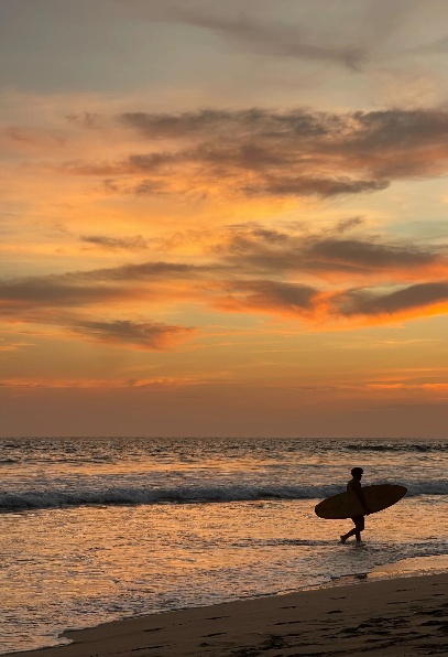 A person carrying a surfboard walking along the beach at sunset, with colorful clouds in the sky and the ocean waves.
