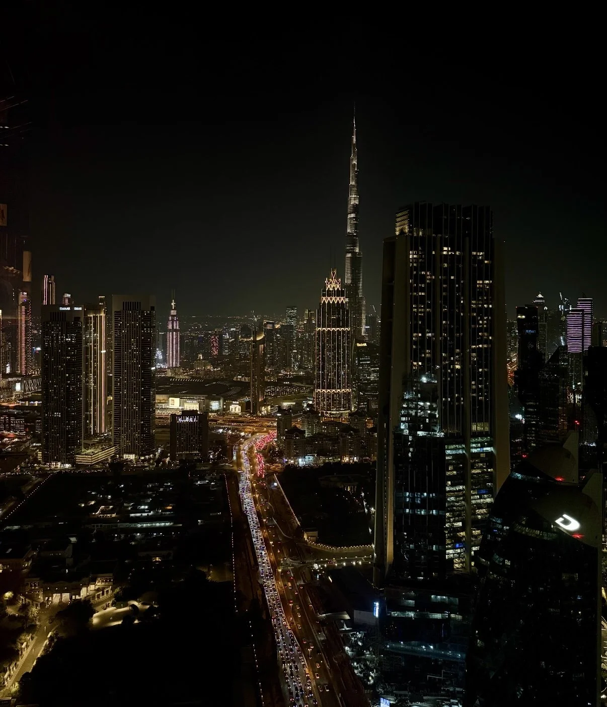 Nighttime cityscape of Dubai with illuminated skyscrapers, including the Burj Khalifa, and busy traffic on a central road.