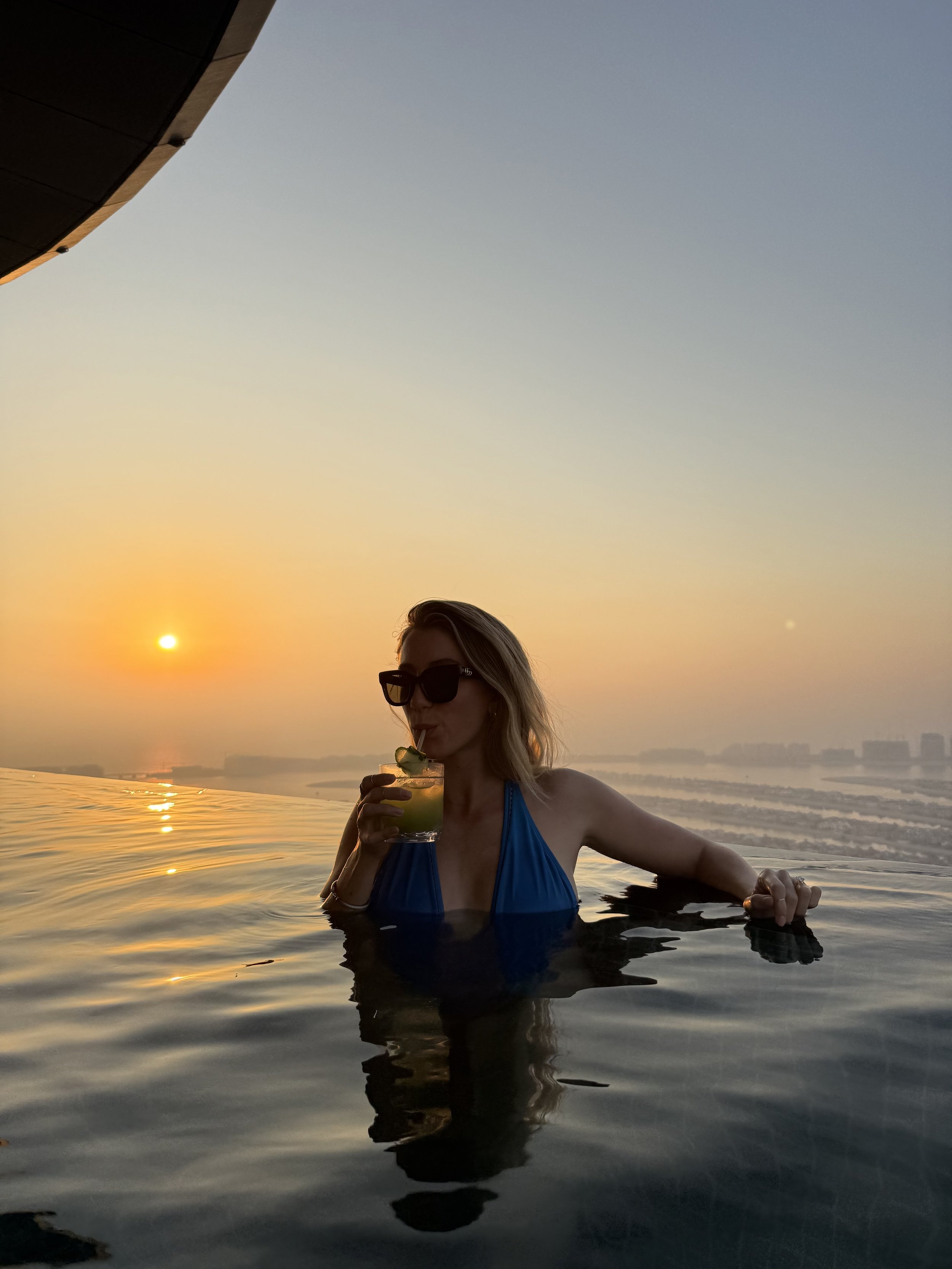 A woman with blonde hair wearing sunglasses and a blue swimsuit, holding a cocktail, relaxing in an infinity pool during sunset.