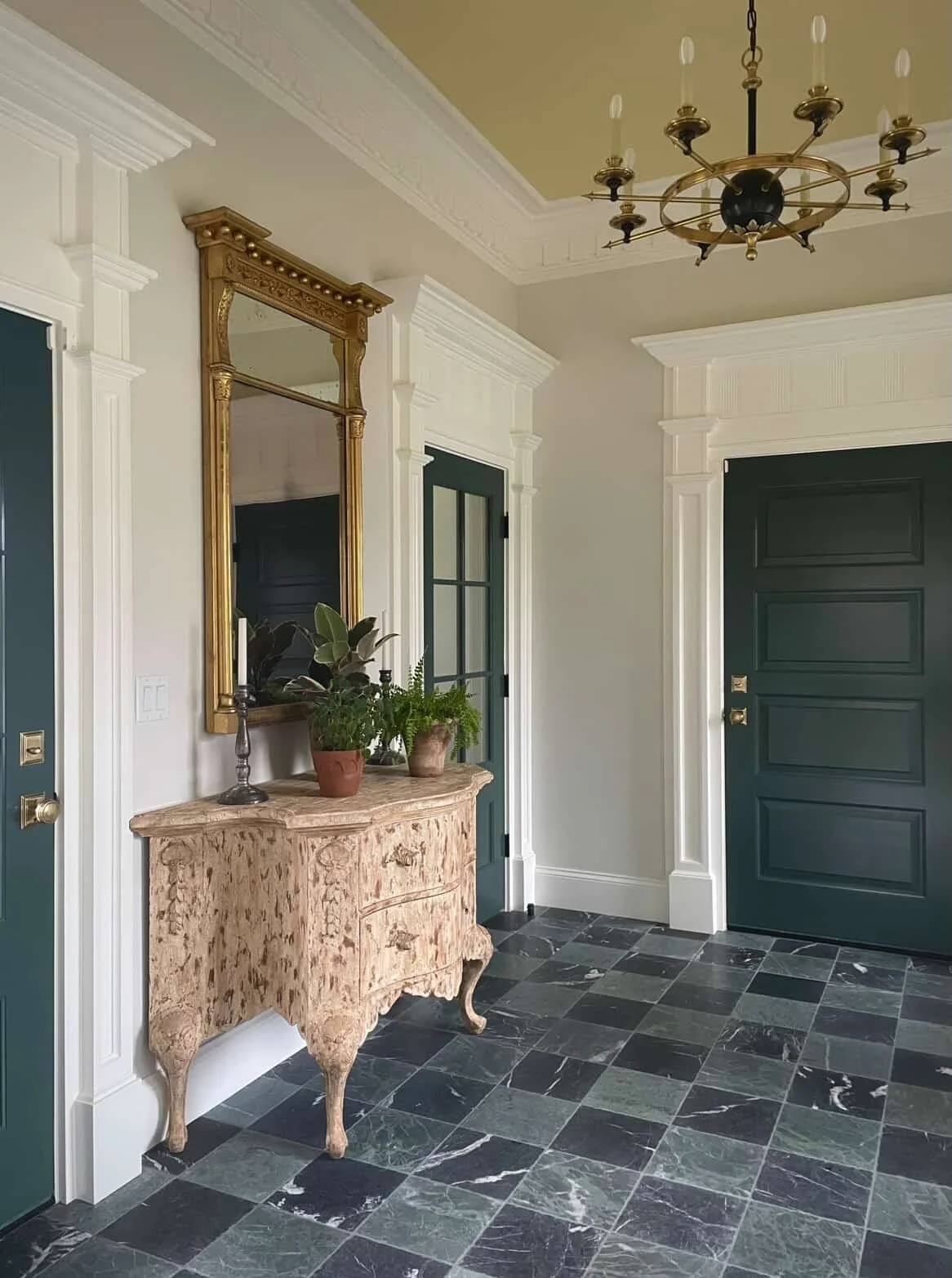 Entryway with dark green door, white decorative trim, marble floor tiles, vintage wooden console table with plants, large gold-framed mirror, and a chandelier hanging from the ceiling.