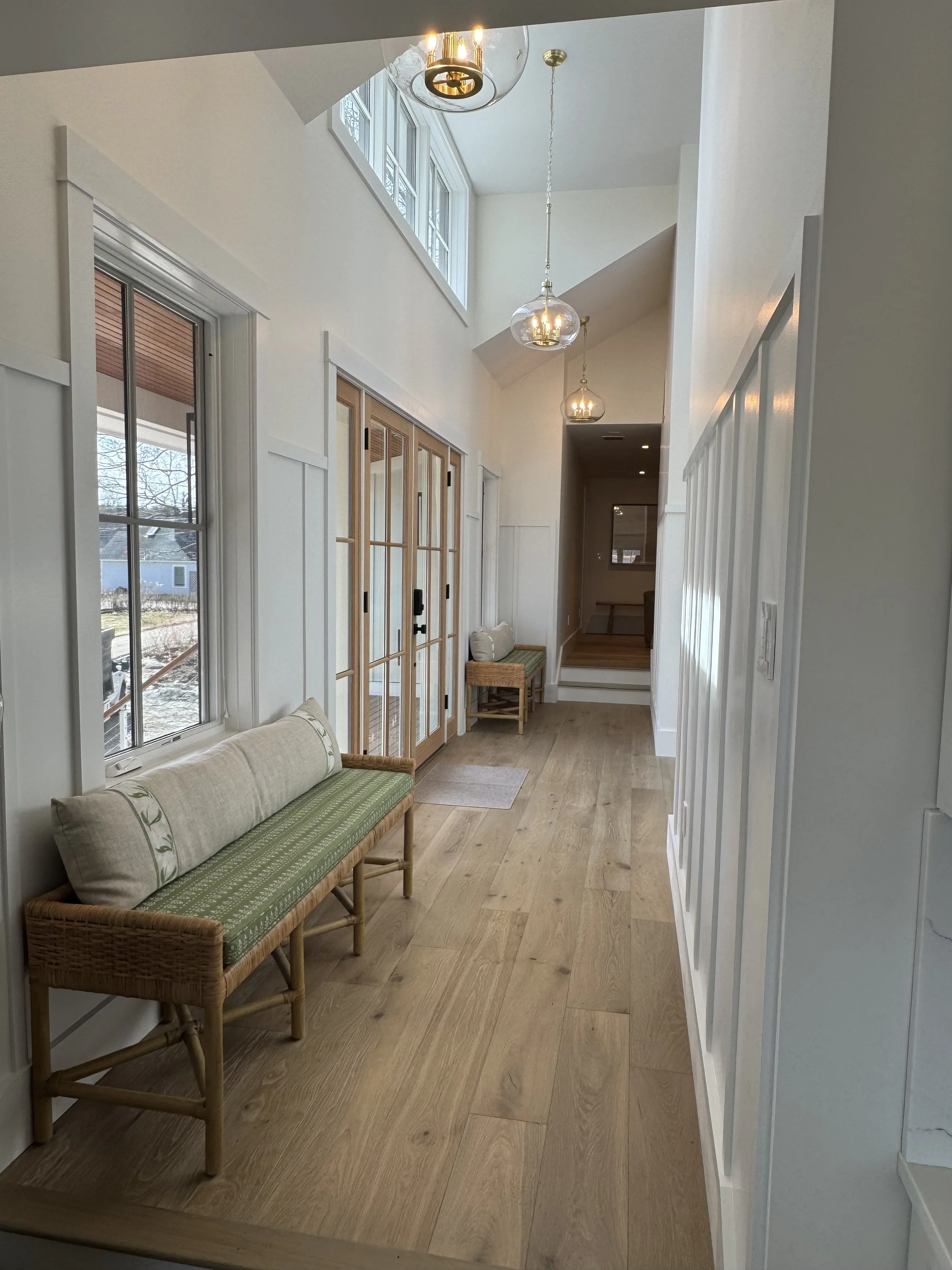 Bright hallway with large windows, wooden flooring, two green cushioned benches, glass pendant lights, and a set of wooden and glass sliding doors.