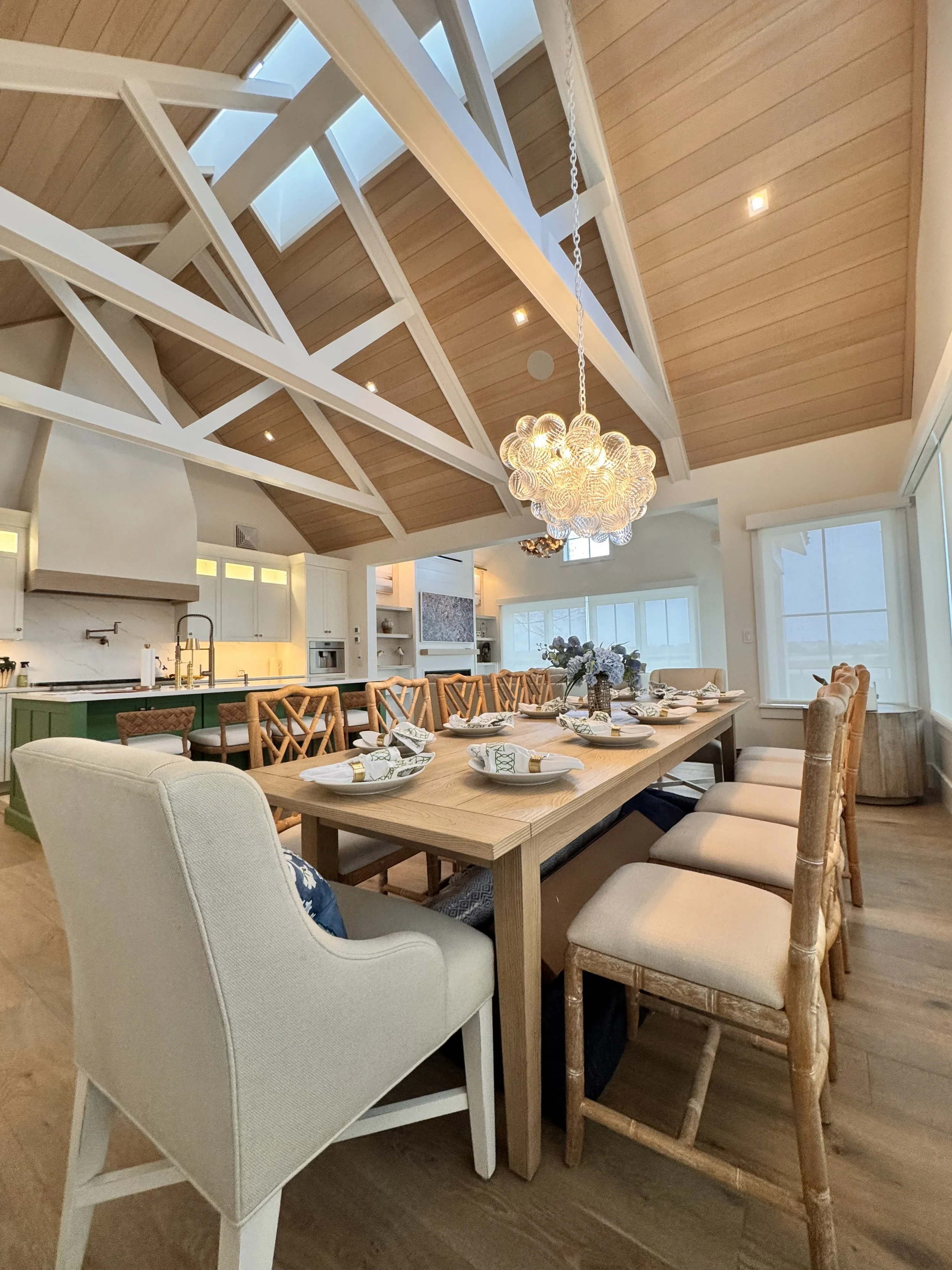 A spacious dining area with a long wooden table set for a meal, surrounded by a mix of upholstered and wooden chairs, under a high vaulted ceiling with exposed beams and skylights, illuminated by a modern chandelier.