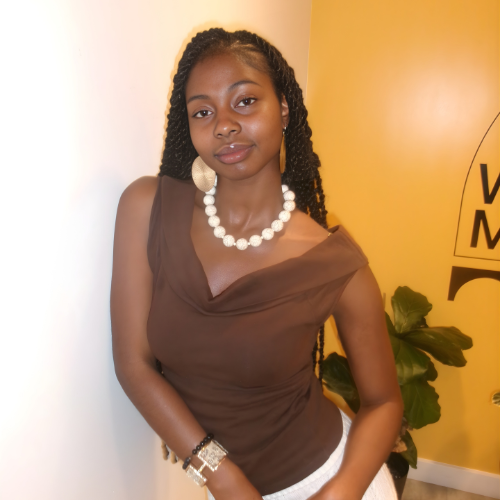 A young woman with braided hair wearing a brown top, a white beaded necklace, earrings, and a bracelet, standing indoors against a yellow wall with a plant nearby.