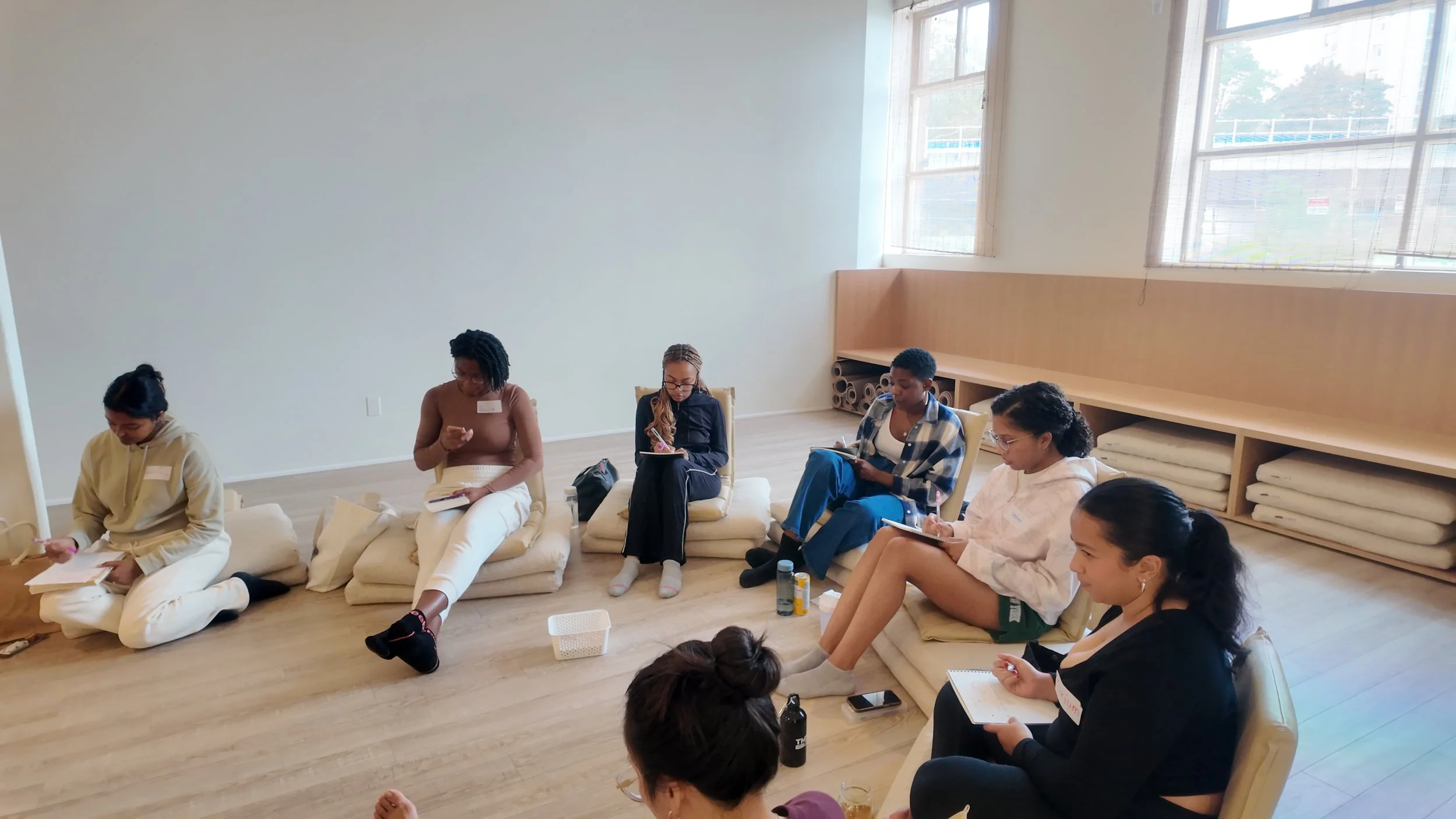 Group of women sitting on floor cushions in a circle during a workshop or class in a bright room with large windows.
