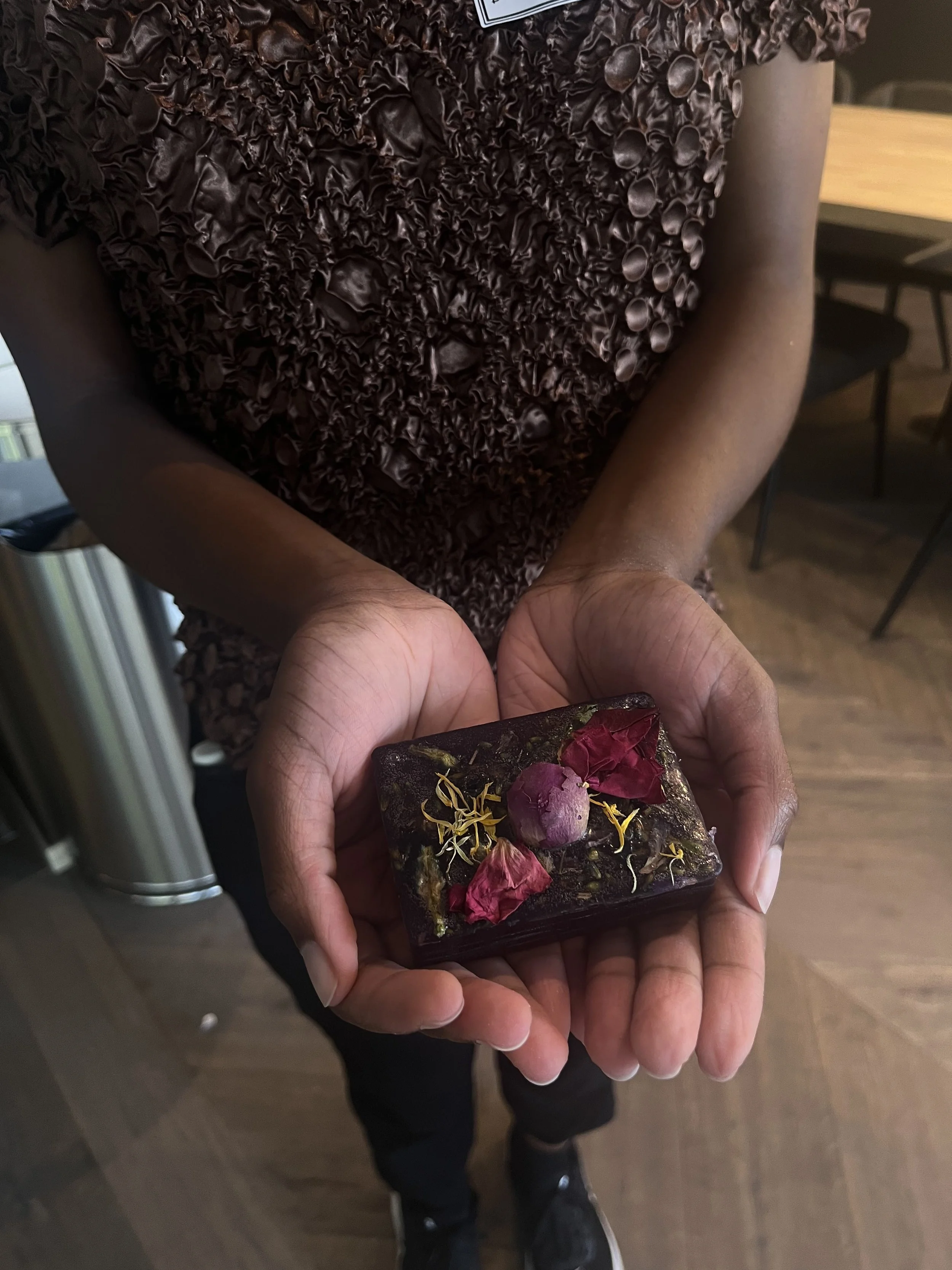 A person holding a decorative rectangular soap or ornament with dried flower petals and a round pinkish element on top.
