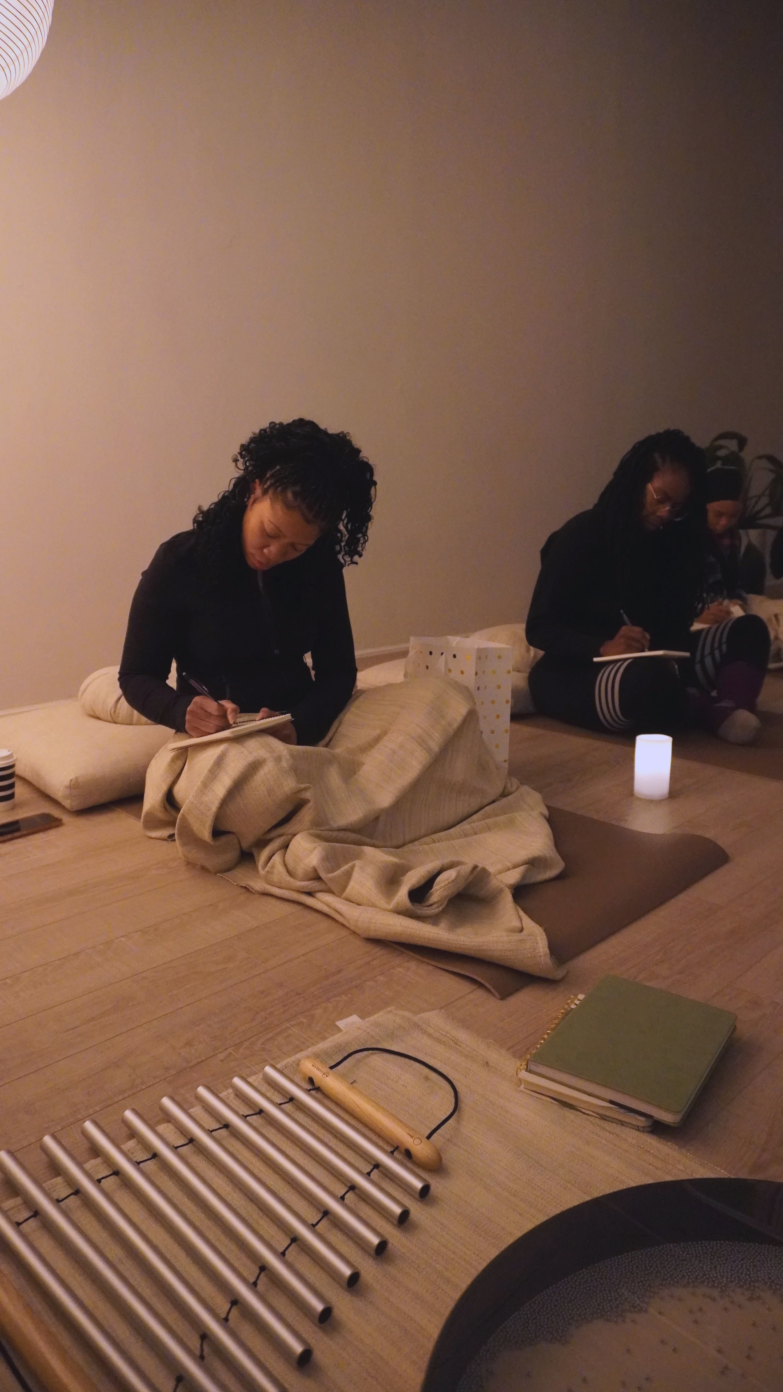two women of colour sitting on cushions on the floor, taking notes in a dimly lit room, with candles and notebooks around them, and a singing bowl nearby.