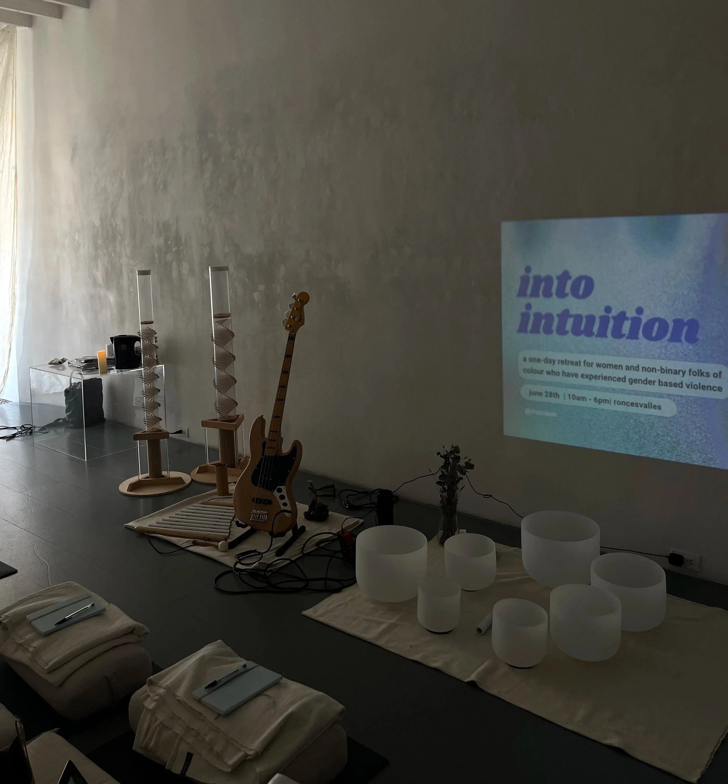 An indoor space set up for a women's and non-binary retreat. There are two wooden torii-style displays, a bass guitar, several white singing bowls of varying sizes on beige mats, and a projector screen displaying an event titled 'into intuition'. The room has a neutral wall, natural light, and table settings with notebooks and pens.