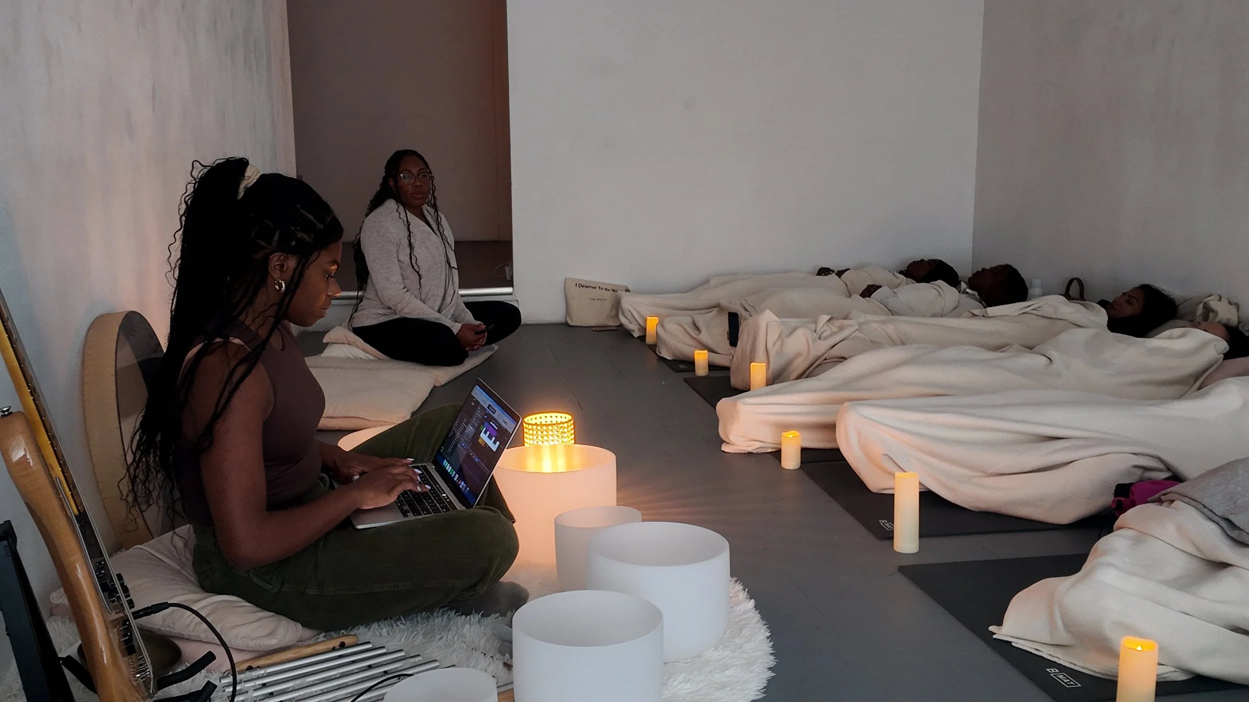 A meditation or yoga session with three women practicing in a dimly lit room. Two women are lying down on mats, covered with white blankets, and holding their own posture and eyes closed. The third woman is sitting cross-legged with a laptop and a laptop light, possibly leading or guiding the session. The room has lit candles and minimalist decor, creating a calming atmosphere.