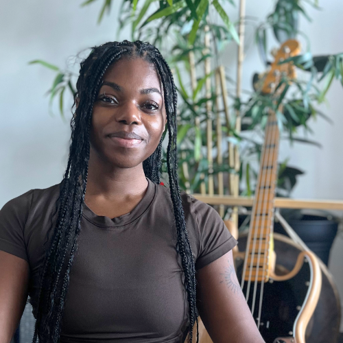 A woman with long braided hair sitting in front of leafy plants and a guitar.