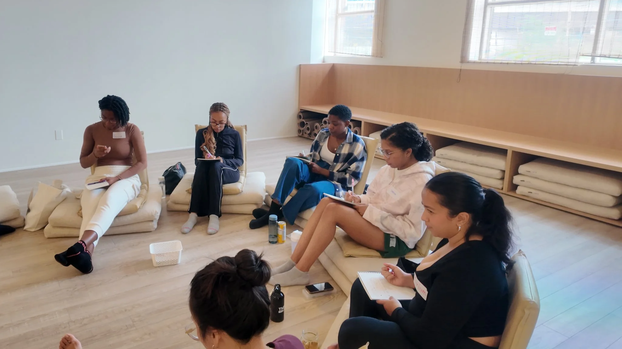 Group of women sitting on cushions in a circle in a bright room, taking notes and listening attentively.